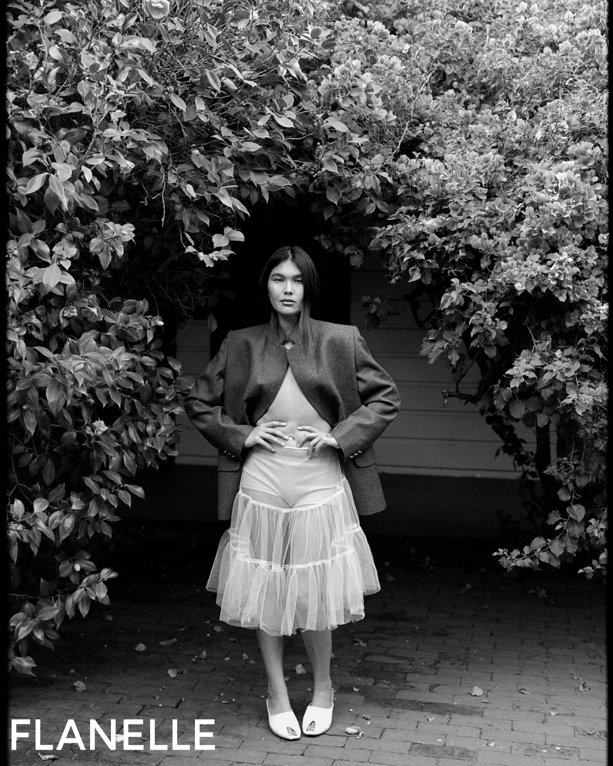 Fashion model in trendy outfit standing under archway of lush greenery.