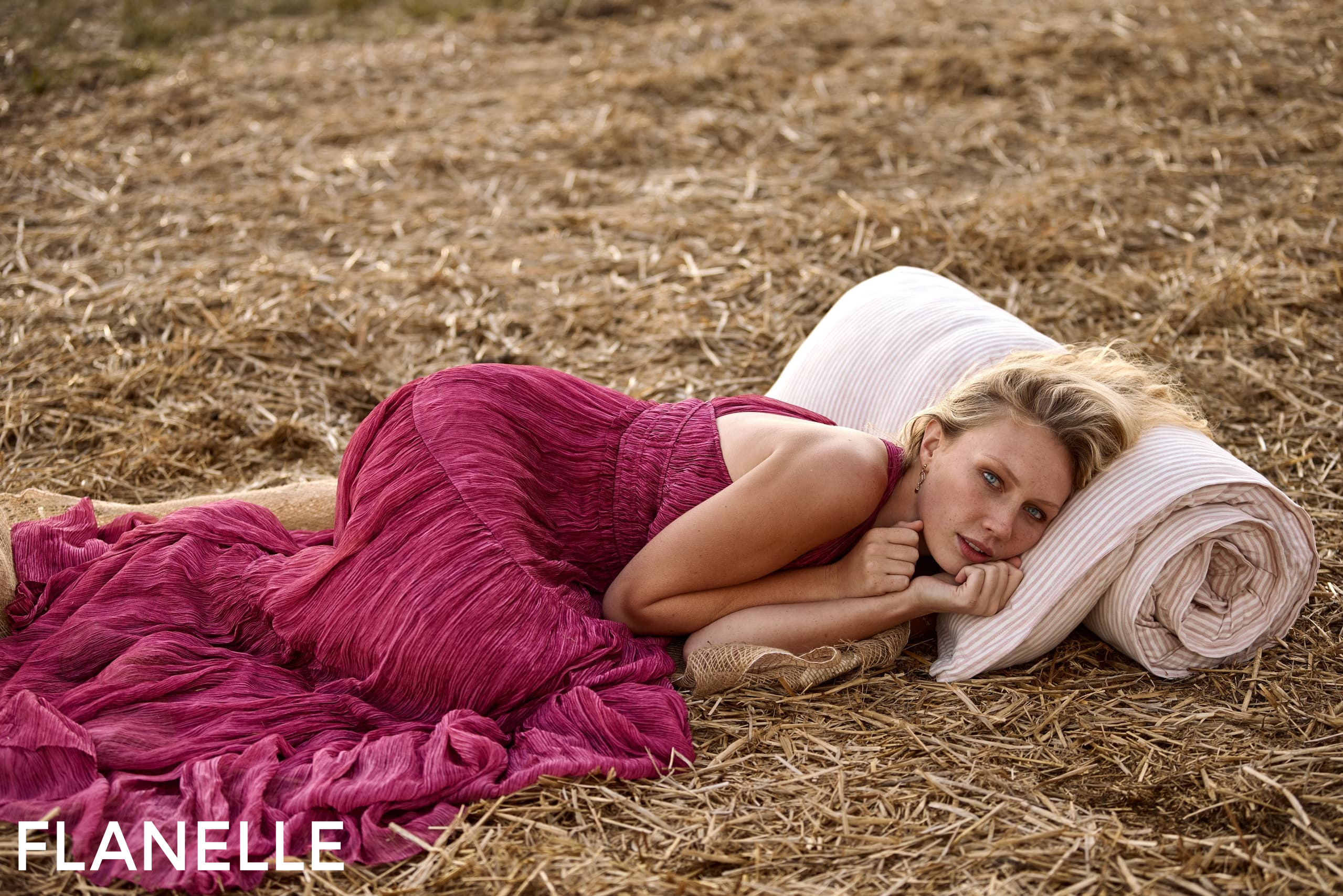 Woman in a pink dress lying on straw with a pillow, serene outdoor setting.