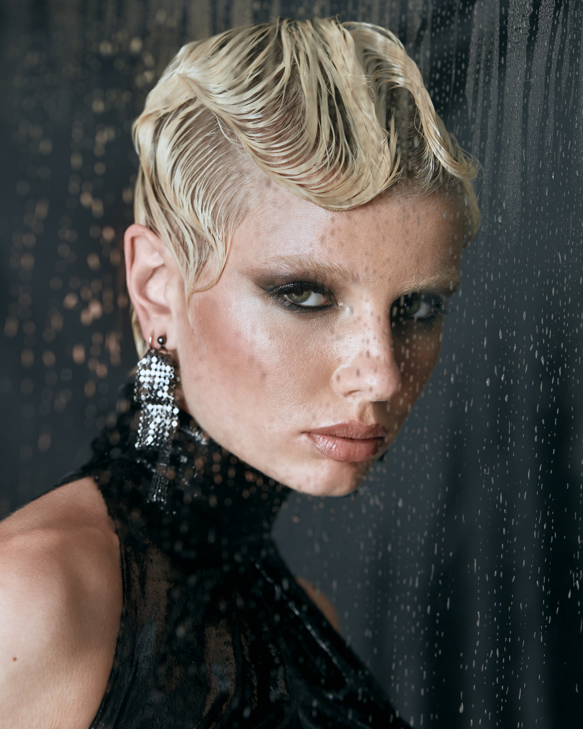 Elegant woman with wet-look hairstyle behind glass with raindrops.