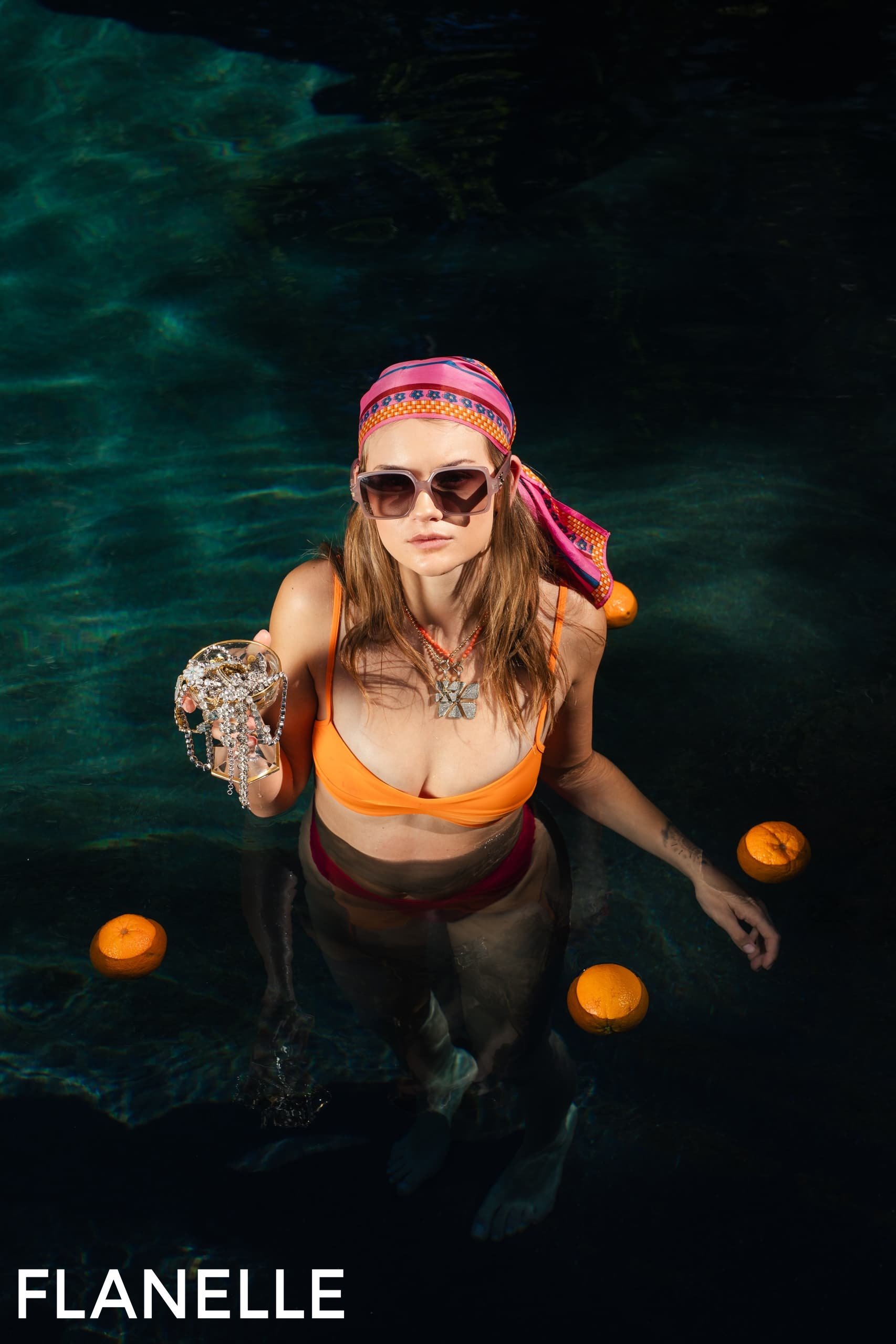 Woman in pool with jewelry, wearing an orange bikini, sunglasses, and headscarf.
