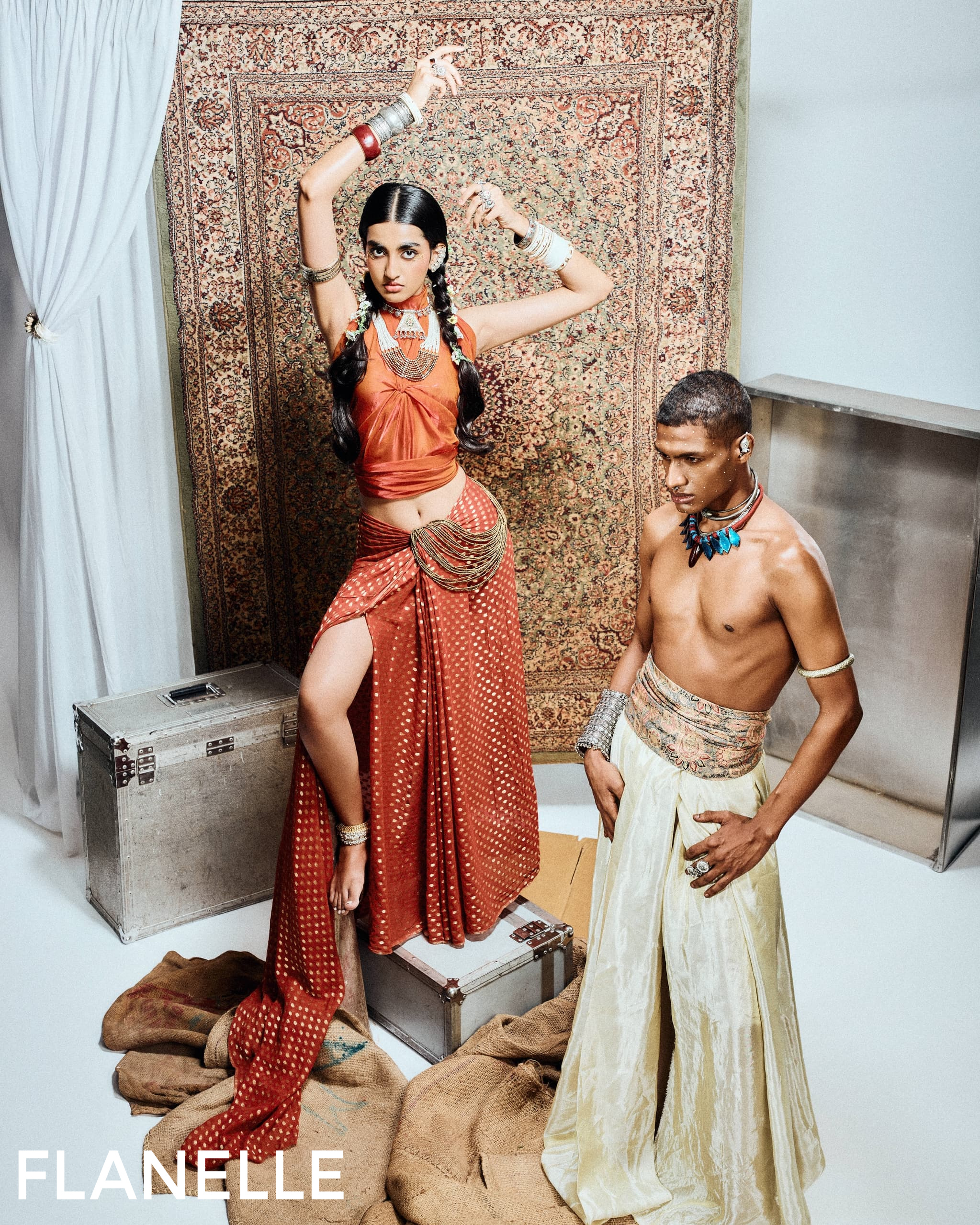 Two models in vibrant attire pose with a patterned rug backdrop.