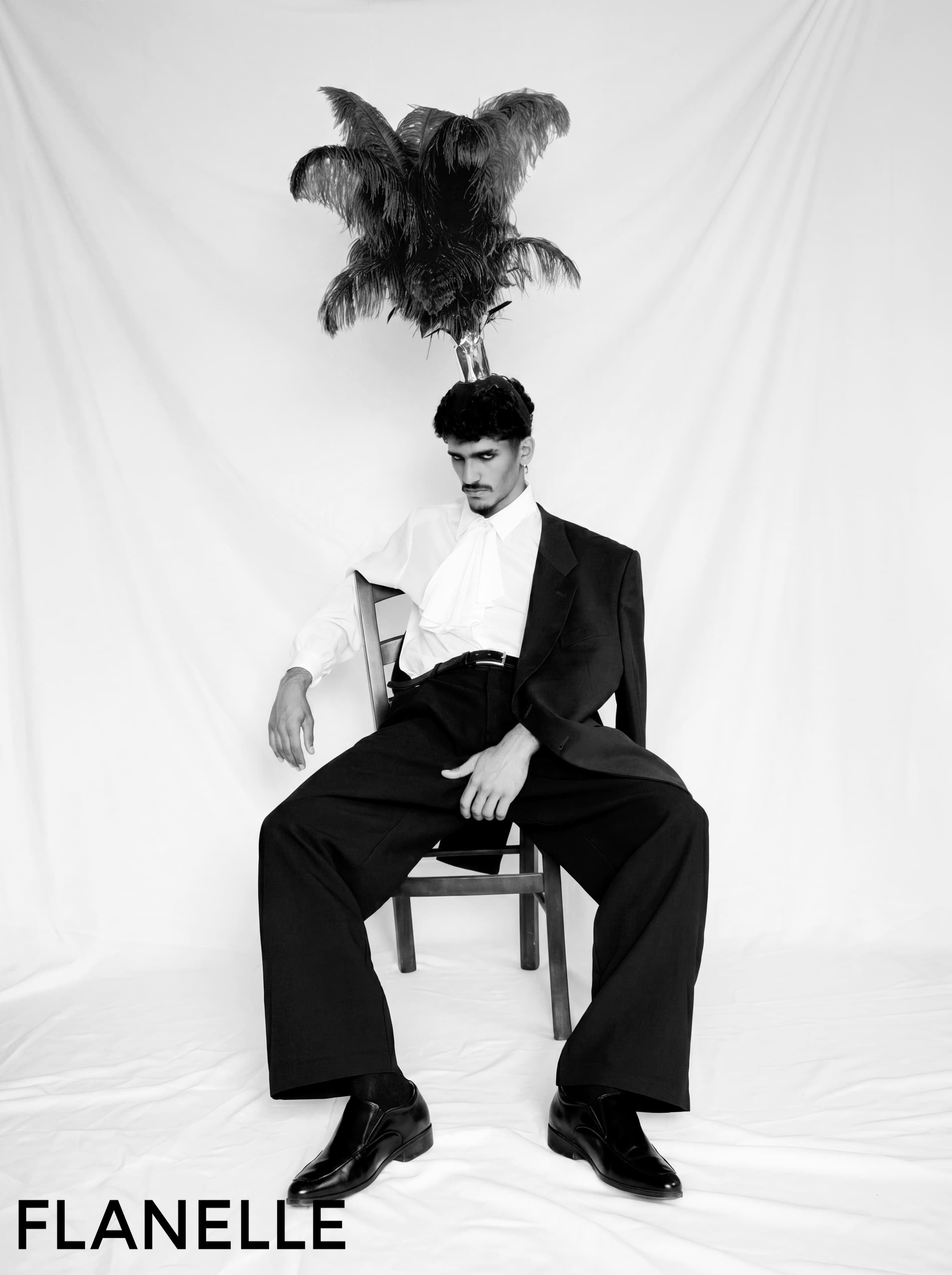 Stylish man in black suit with feathers on head, seated on chair. Fashion photography.