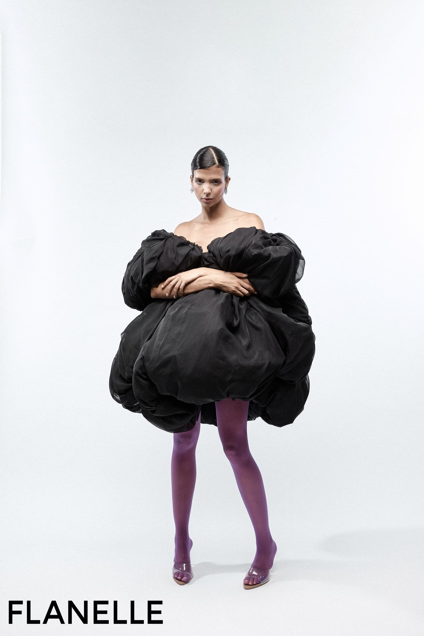 Fashion model in a dramatic black dress and purple tights on a minimalist background.