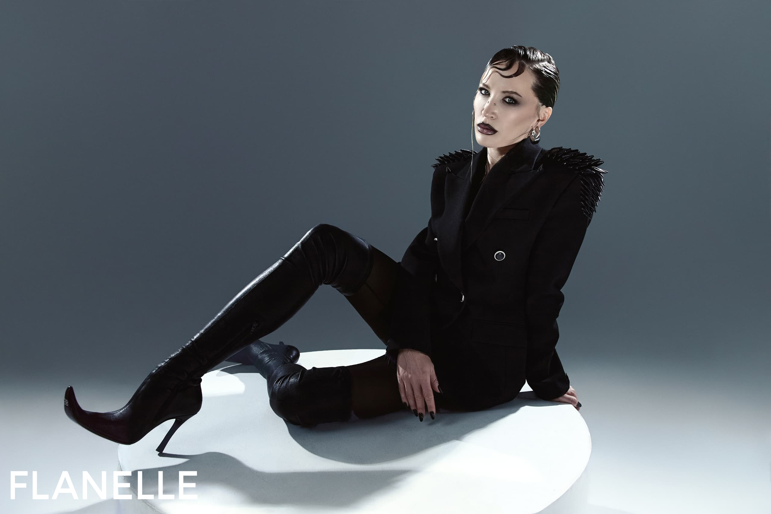 Fashion model in edgy black outfit and boots posed on a white surface.