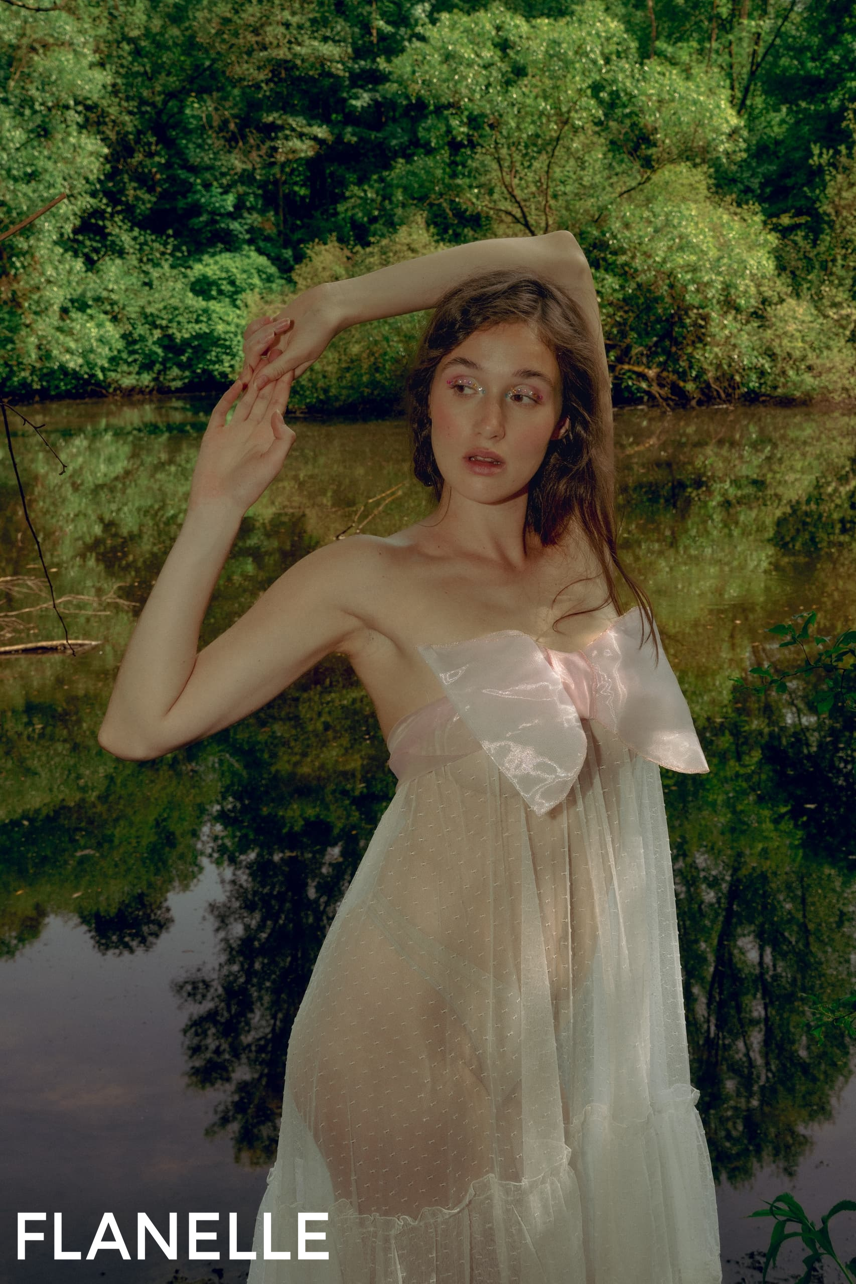 Model in sheer dress poses by a serene pond in lush greenery.