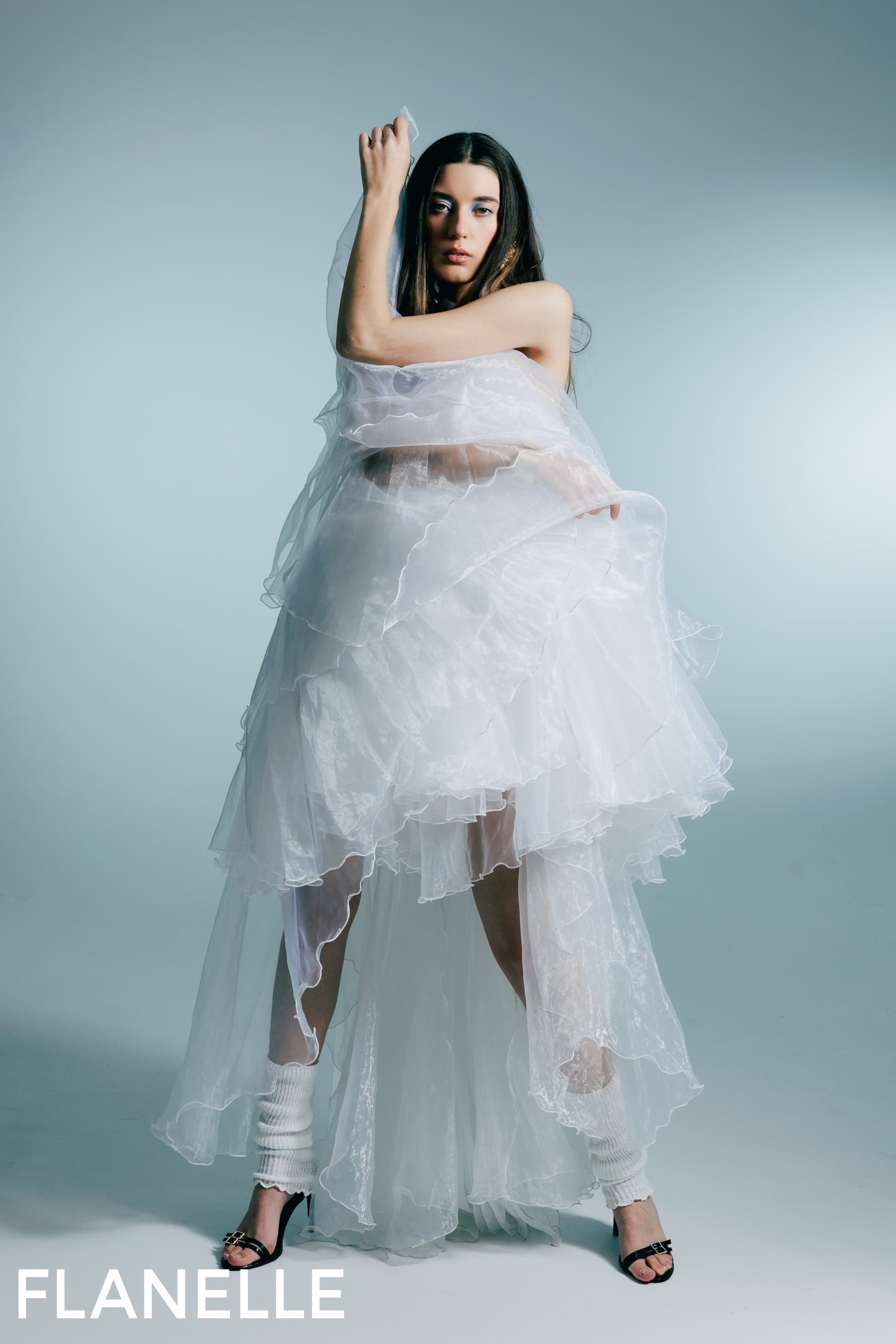 Model posing in layered white dress with sheer fabric against a neutral backdrop.