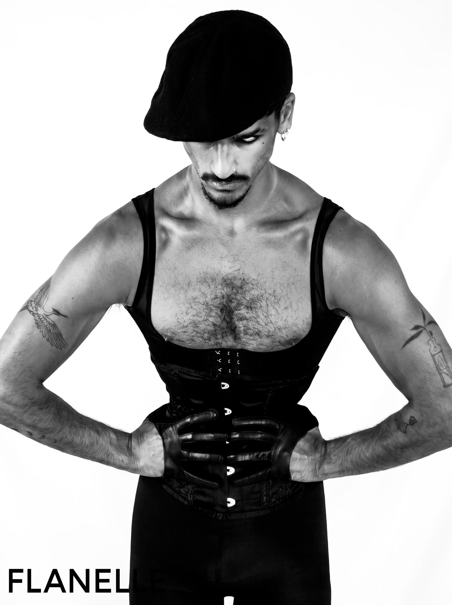 Man in black corset and cap, showcasing tattoos and gloves in edgy fashion photo.