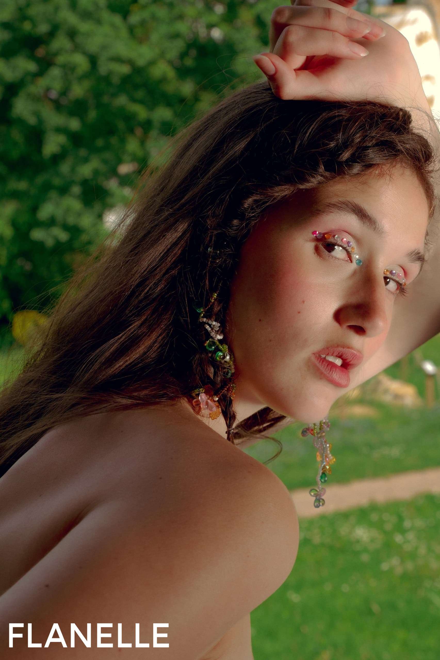 Model with artistic makeup poses outdoors, adorned in colorful jewelry.