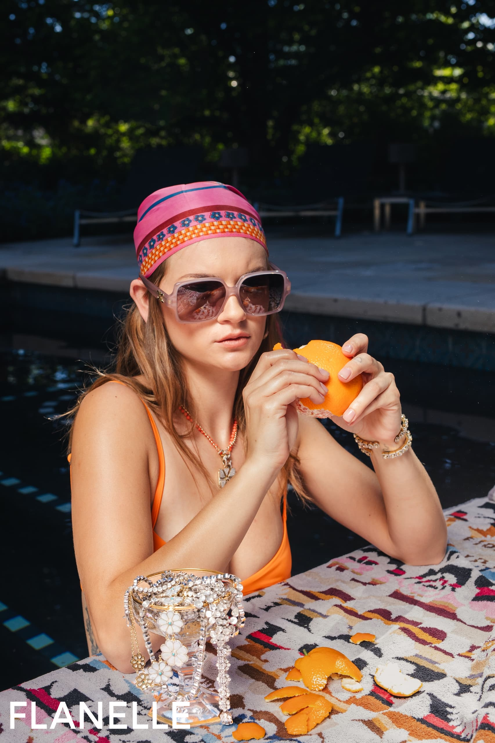 Woman by pool in sunglasses, peeling orange, with jewelry on table. Fashionable scene.