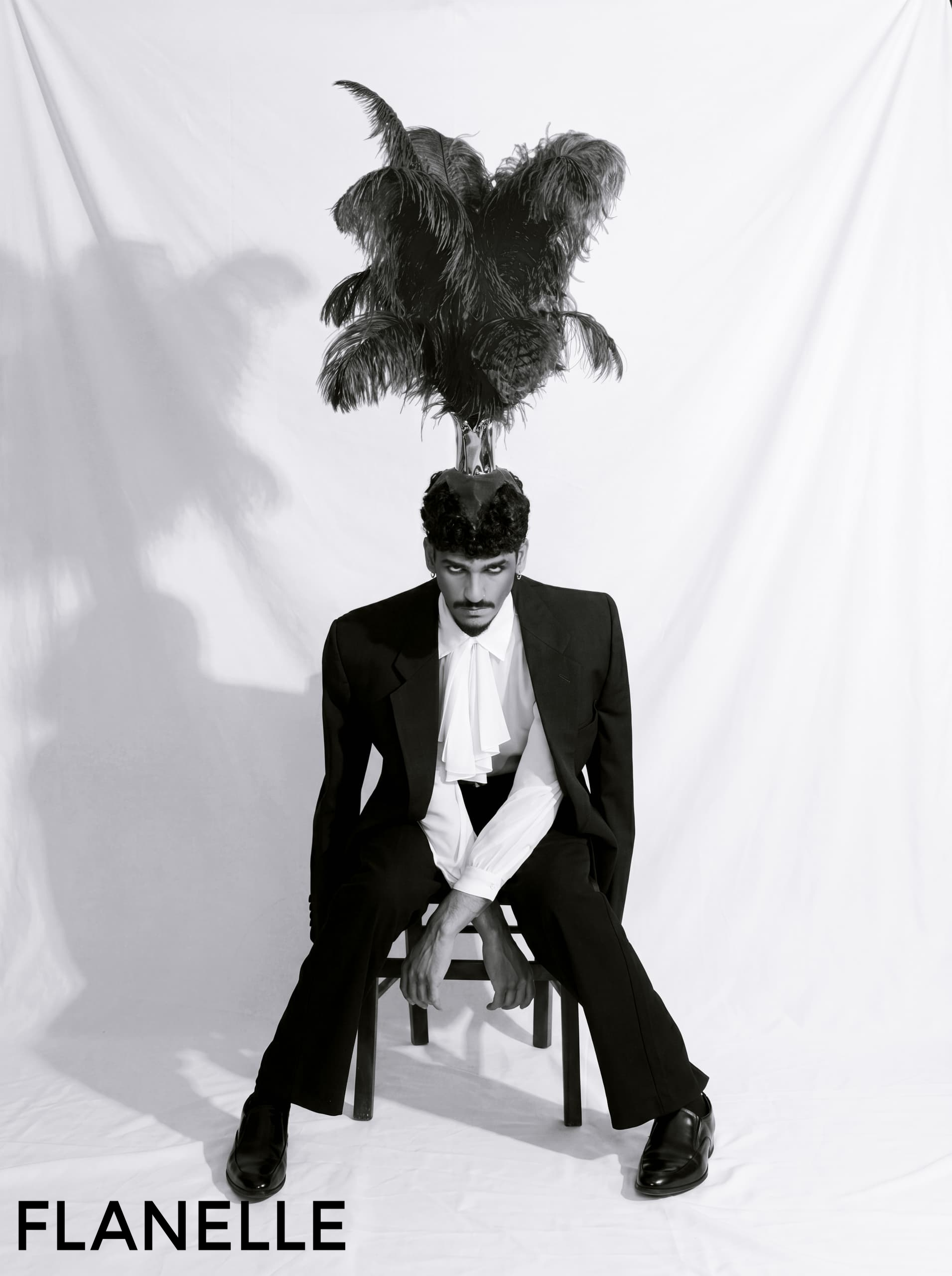 Man in a suit with feathered headpiece, seated against white backdrop.