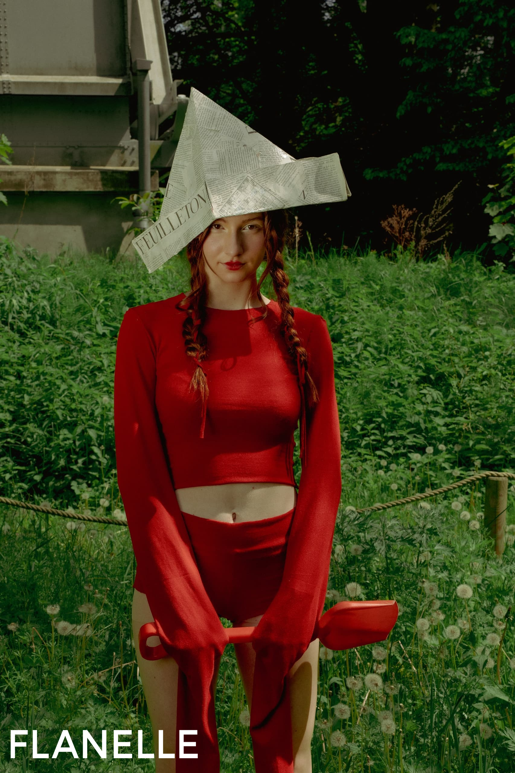 Woman in red with newspaper hat in a lush field.