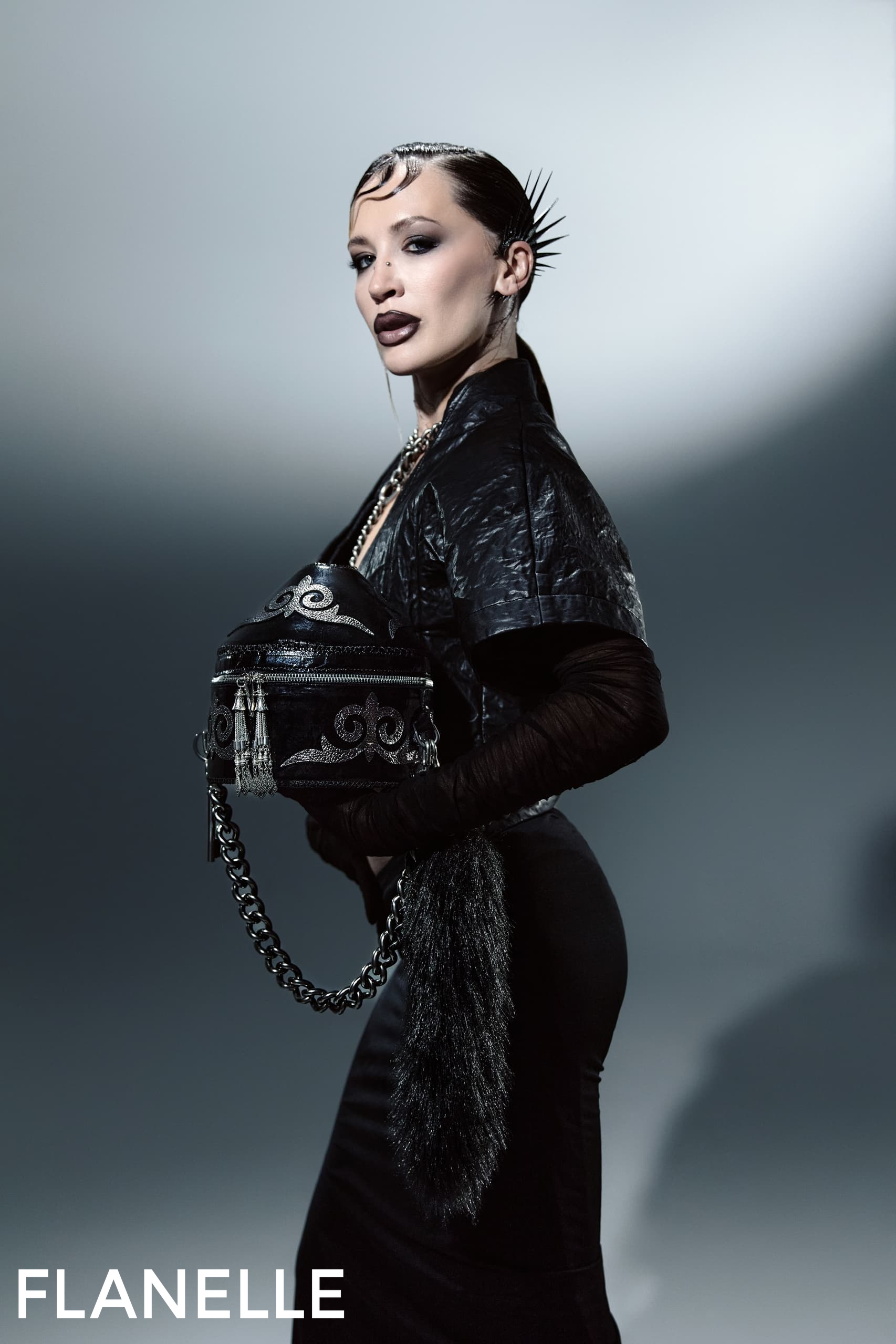 Fashion model in edgy black attire, sleek hair, holding stylish handbag.