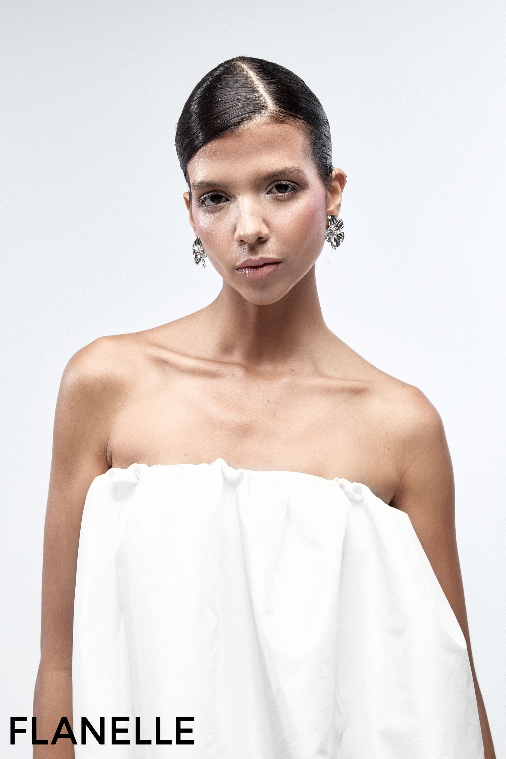Model in elegant white dress with floral earrings and slicked-back hair.