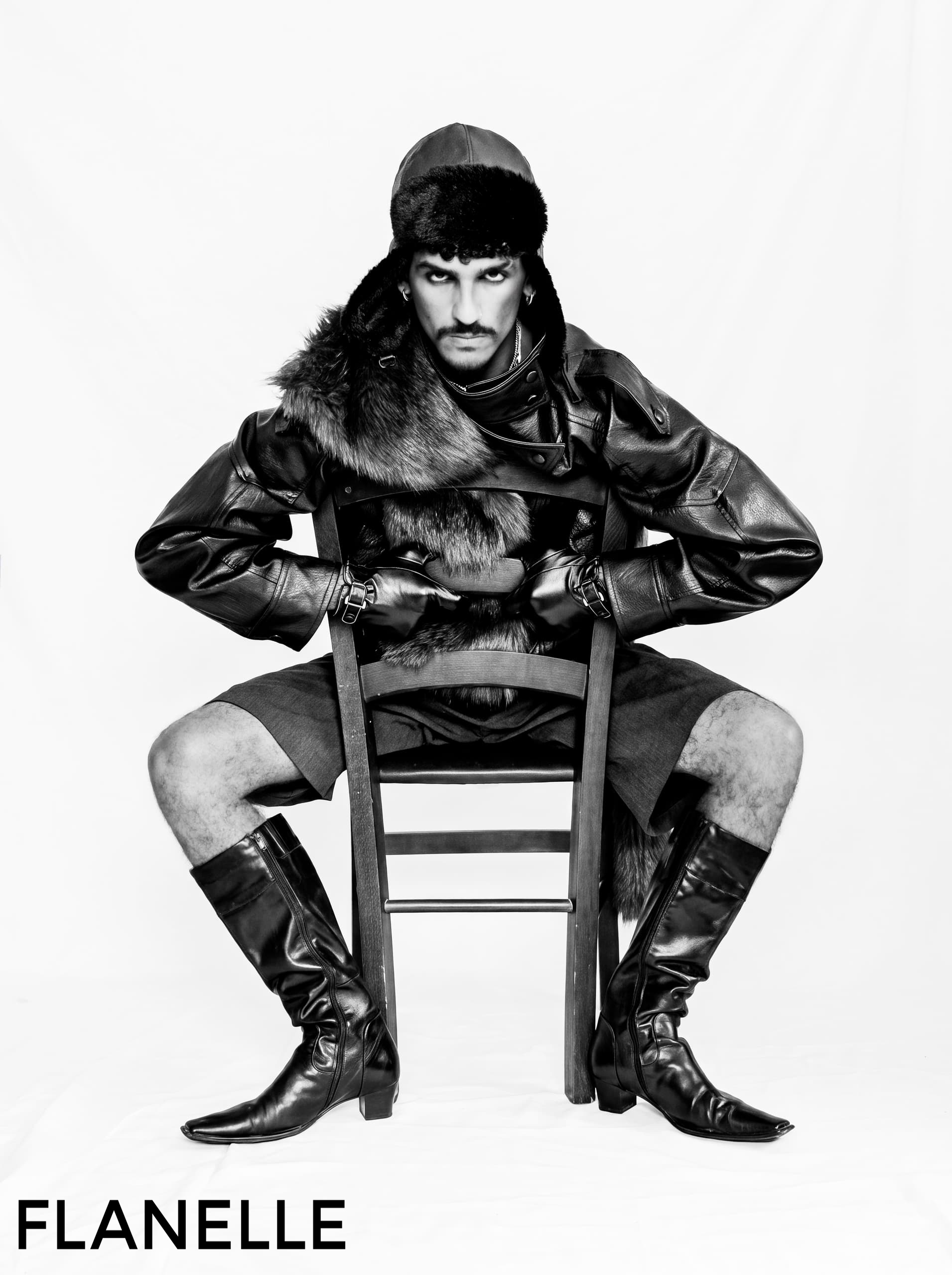 Man in fur and leather, seated on chair, wearing boots. Monochrome fashion editorial.