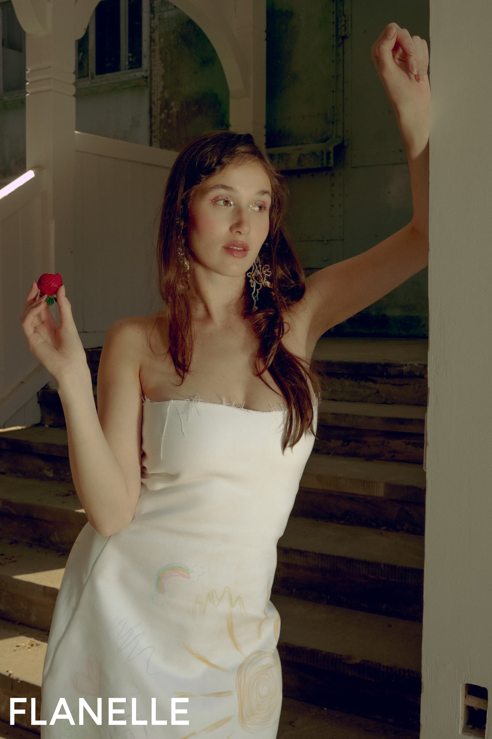 Woman in white dress holding a berry, posed on stairs, branded with FLANELLE.