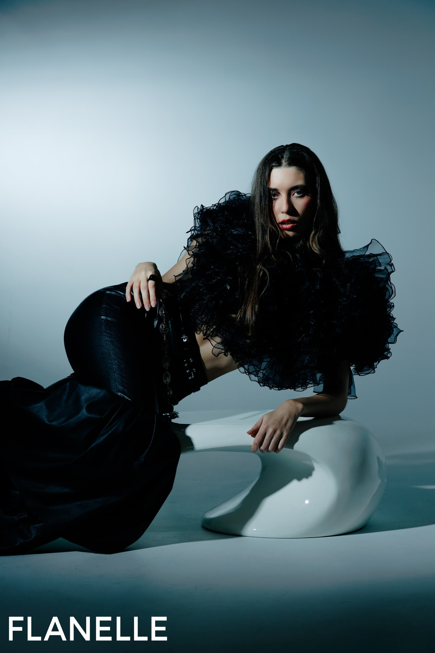 Model in black ruffled outfit poses on sleek modern chair.