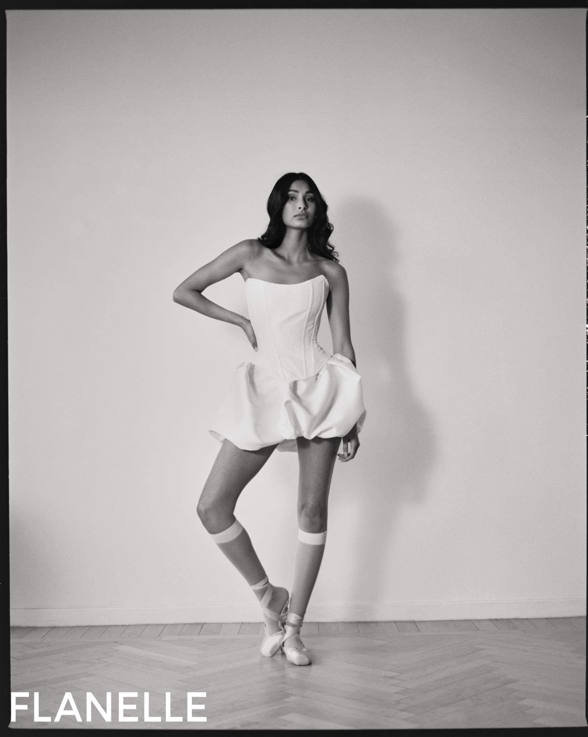 Model in elegant corset dress and ballet shoes poses confidently against a plain backdrop.