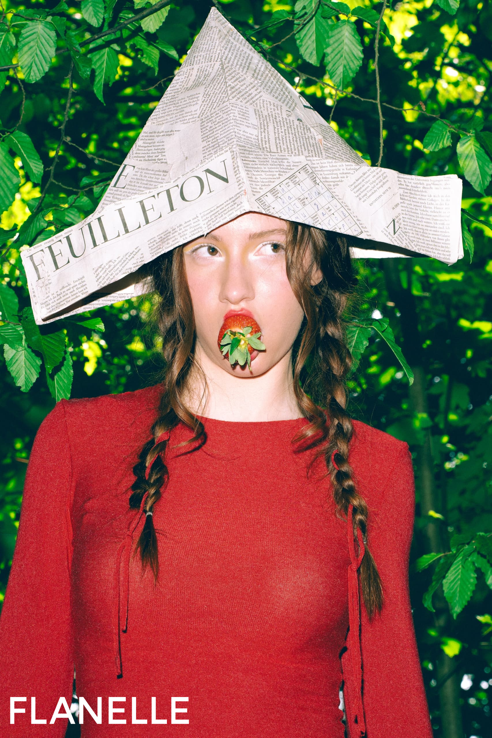 Woman in red with newspaper hat, holding a strawberry in forest setting.