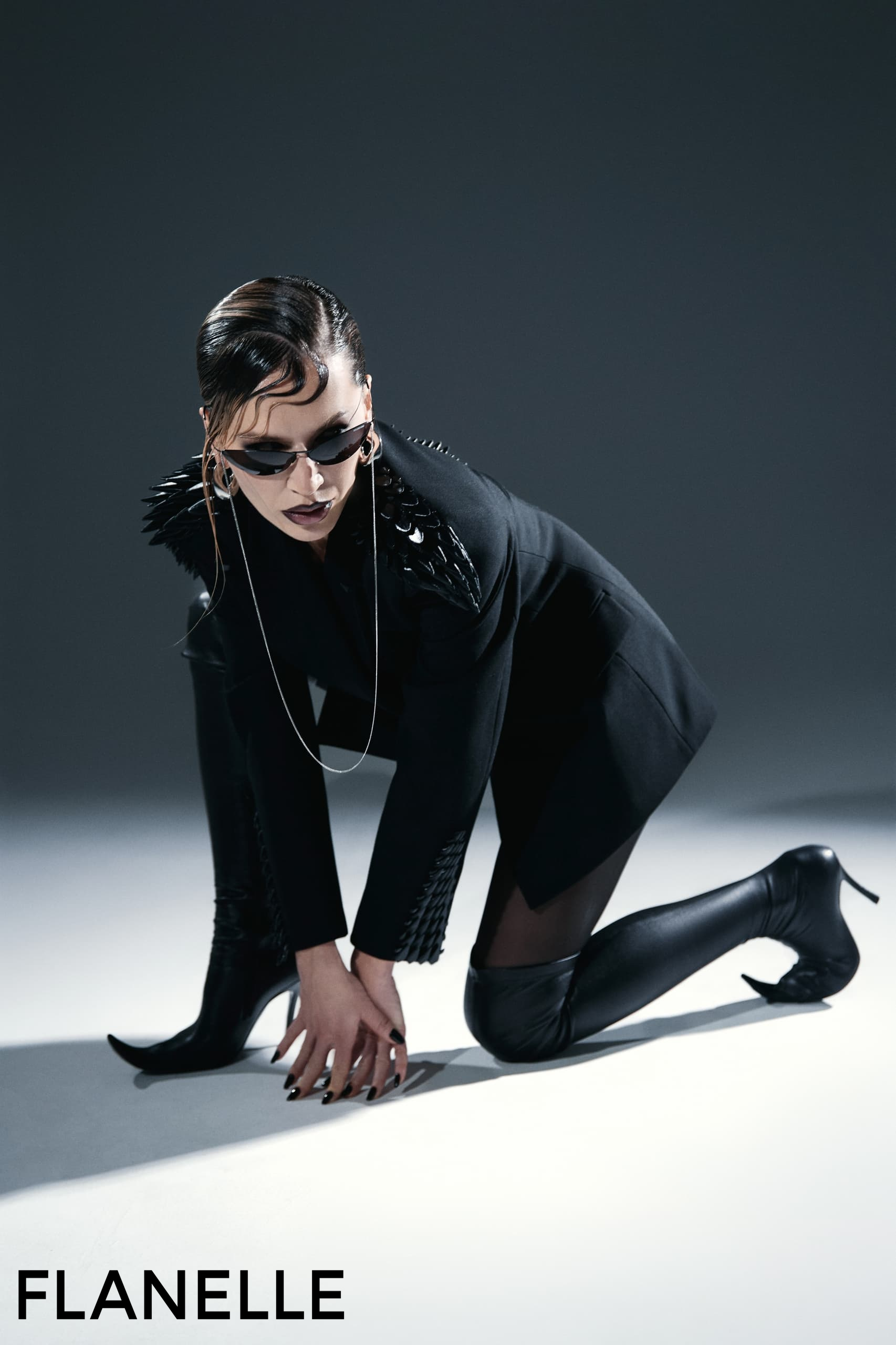 Fashionista in edgy black outfit and shades kneels in a dynamic studio pose.