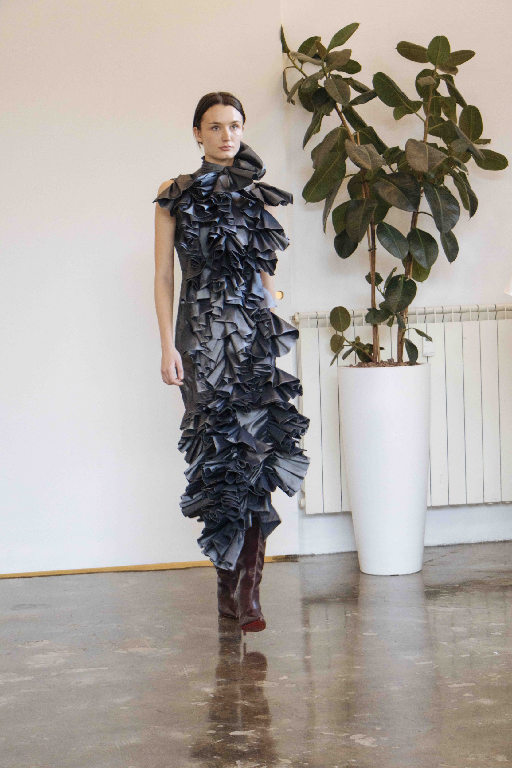 Model in a ruffled dress standing by a plant in a minimalist room.