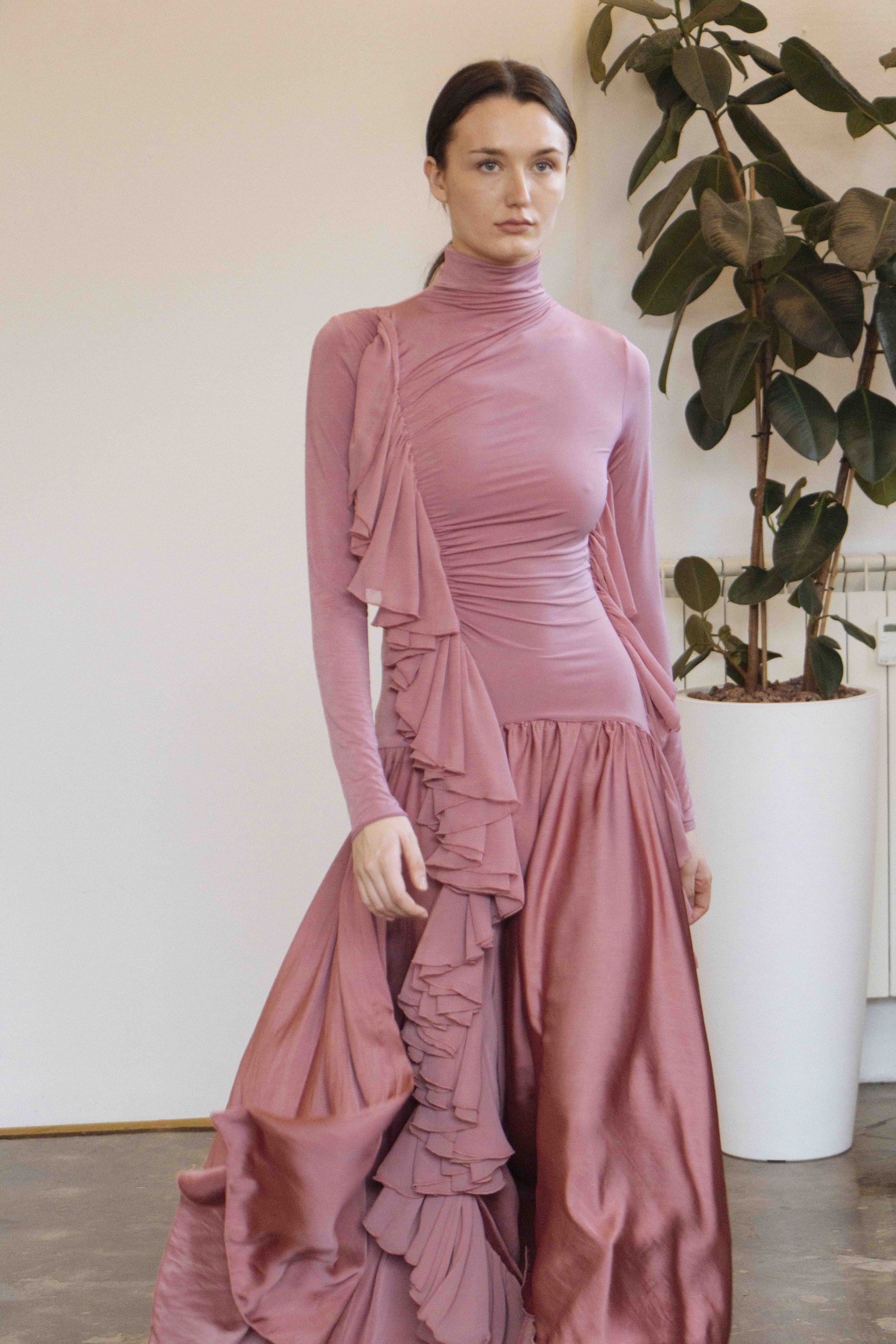 Model in elegant pink dress featuring asymmetrical ruffles, standing indoors by a plant.