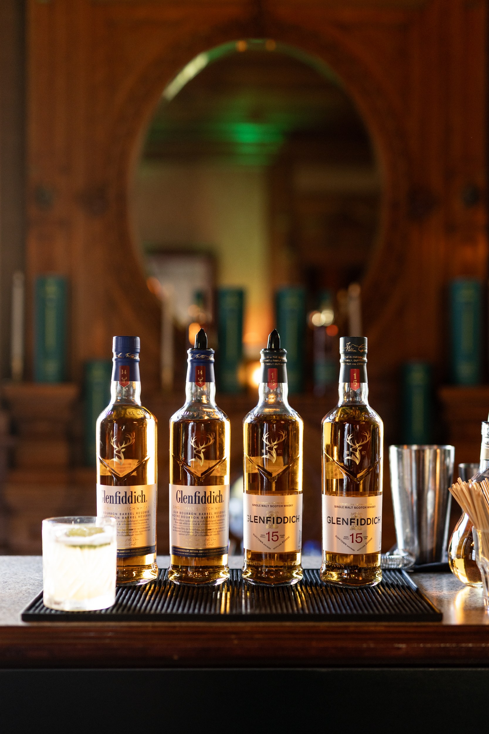 Four Glenfiddich bottles and a drink glass on a bar counter.