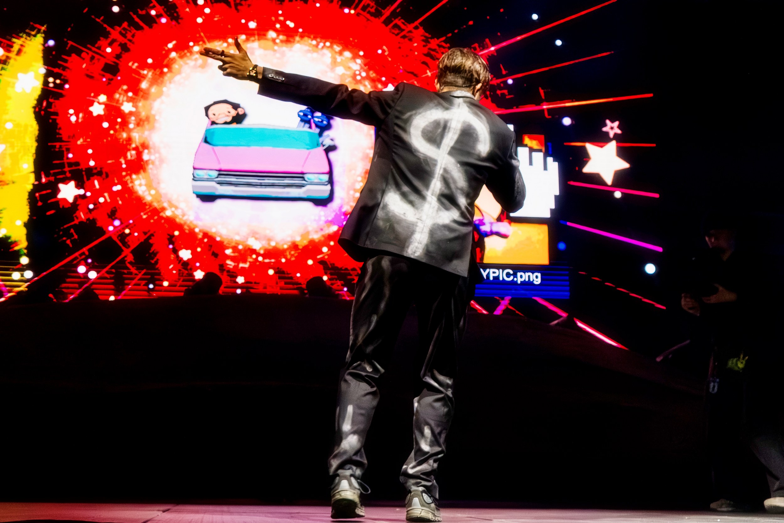 Performer on stage with vibrant digital backdrop and dollar symbol on jacket.