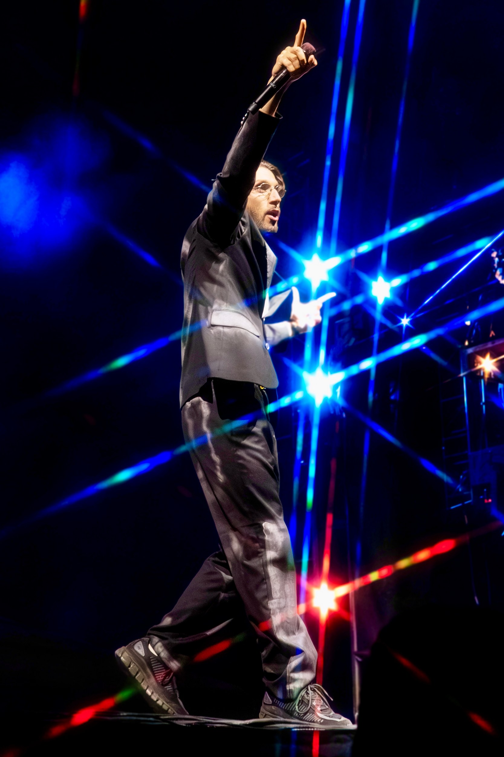 Performer on stage with colorful light effects, holding a microphone and pointing up.