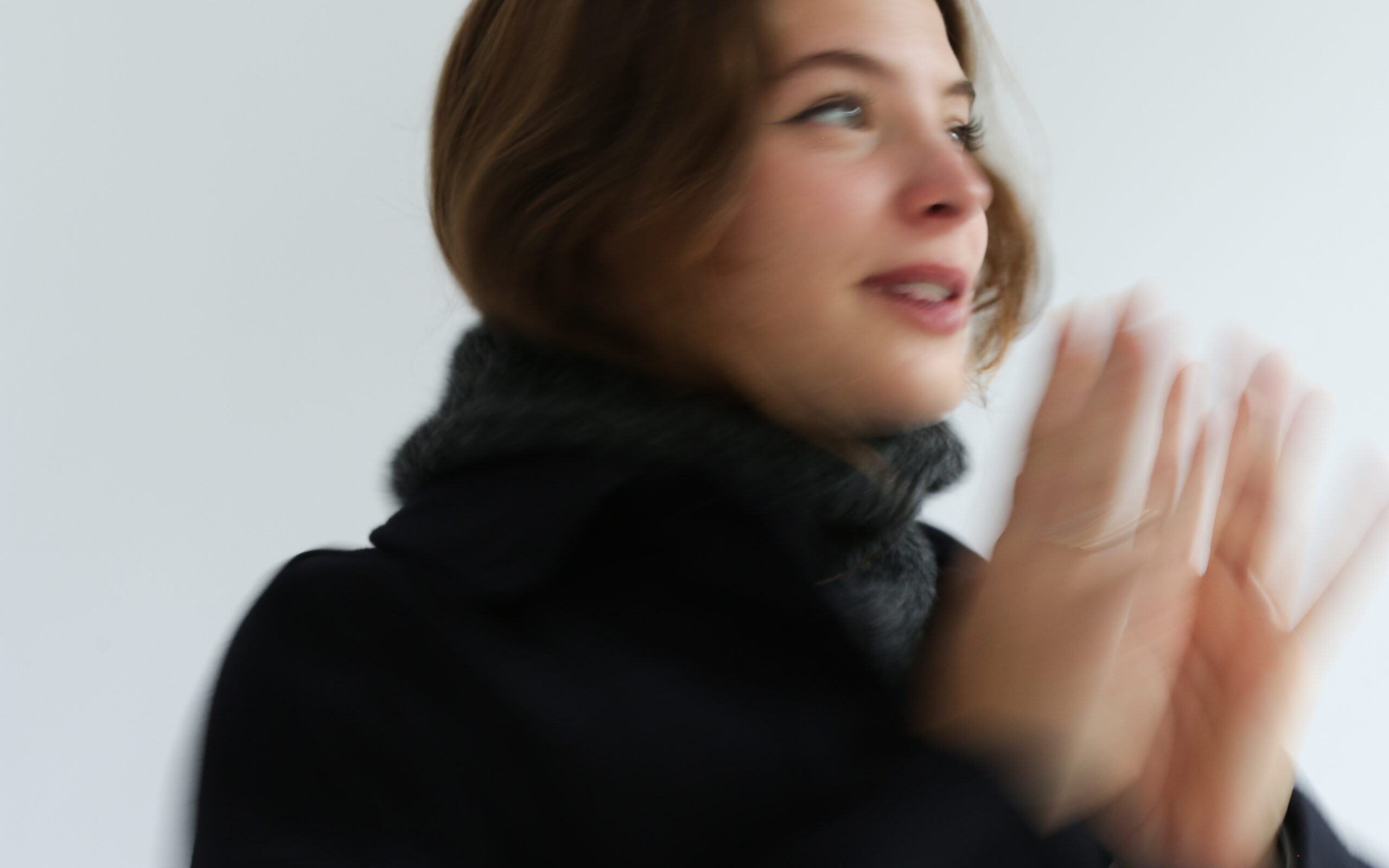 Blurred woman in winter coat smiling, clapping hands, with a neutral background.