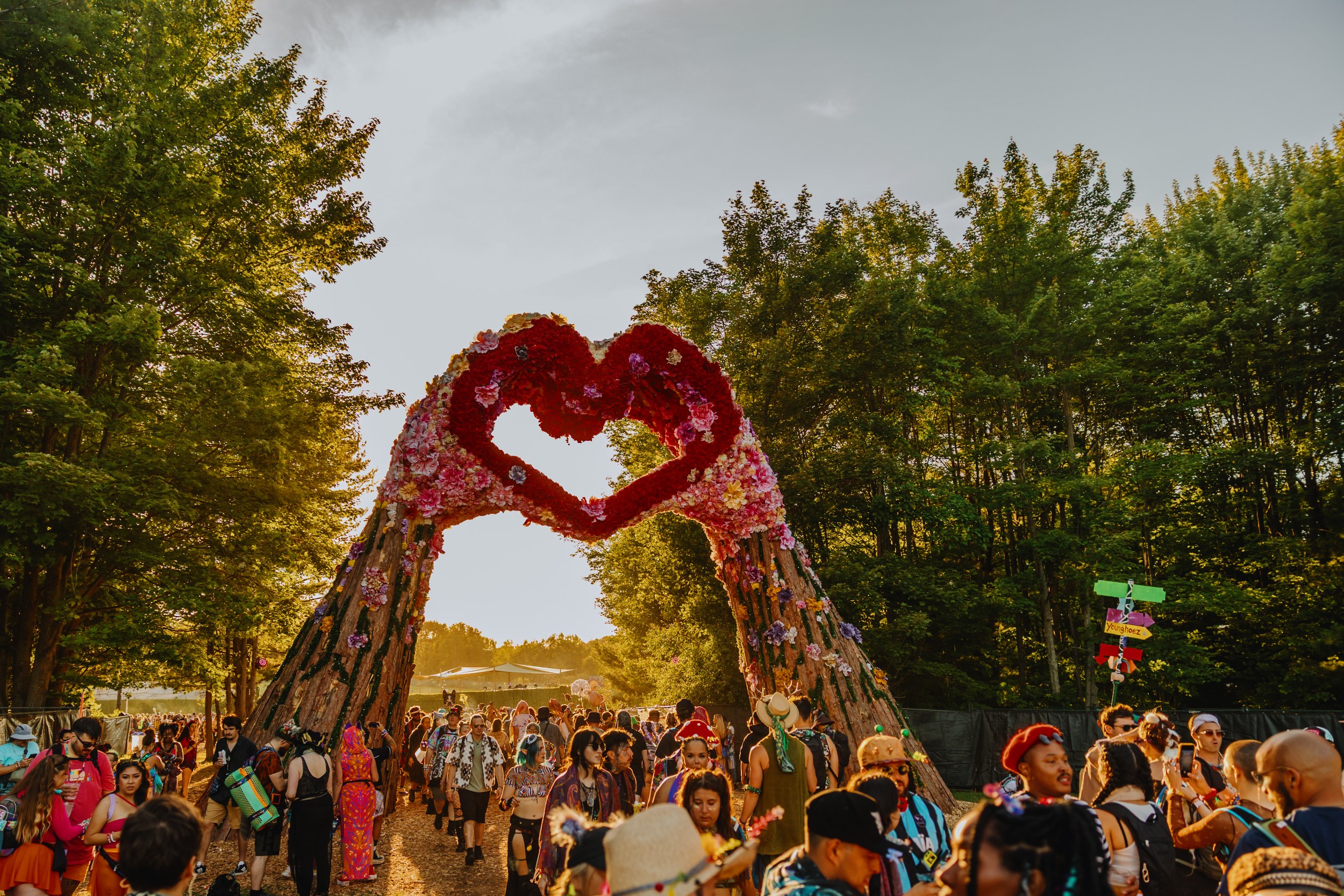 Festival-goers beneath a vibrant heart-shaped floral arch, surrounded by trees.