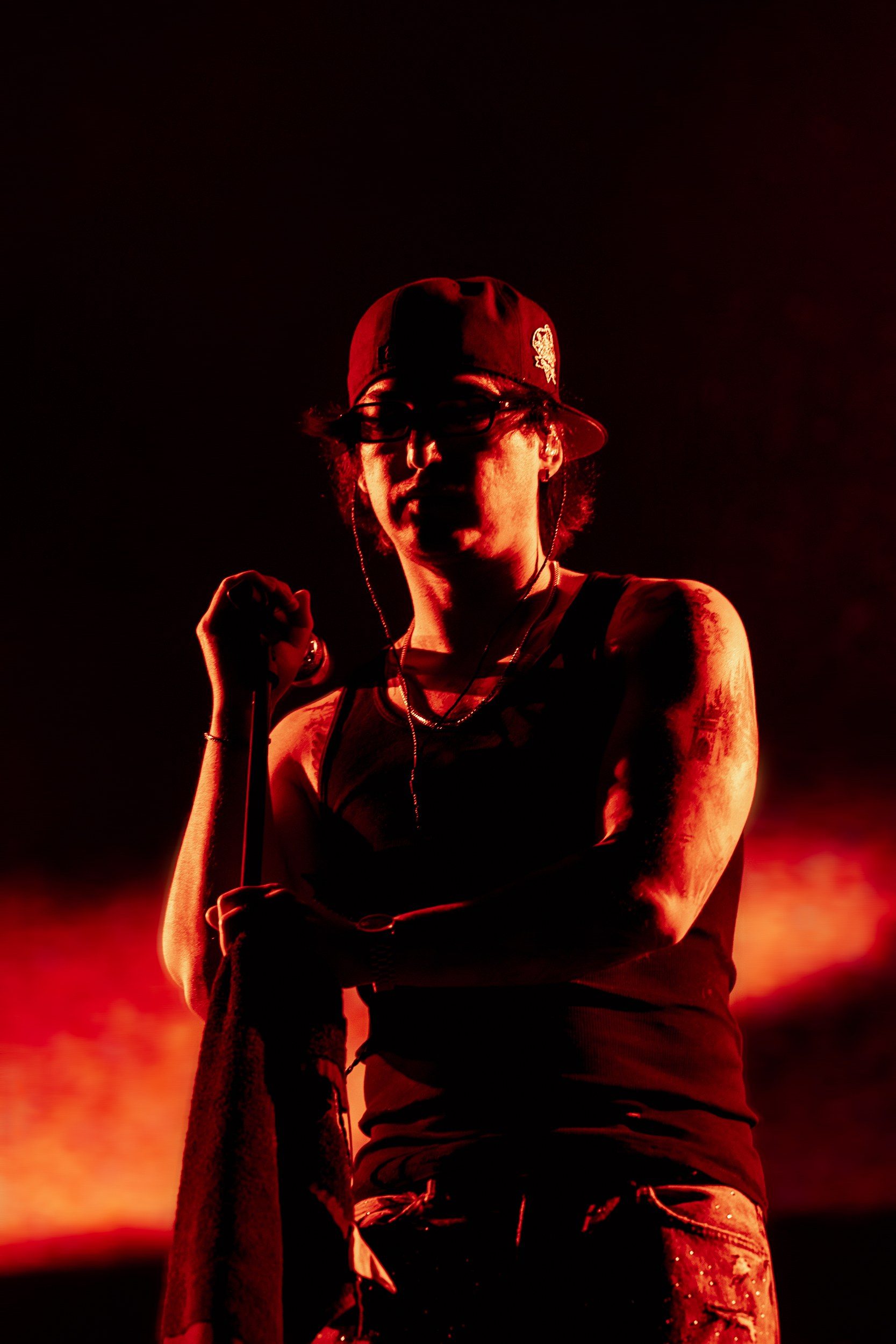 Performer on stage with red lighting, holding a microphone, wearing a cap and tank top.