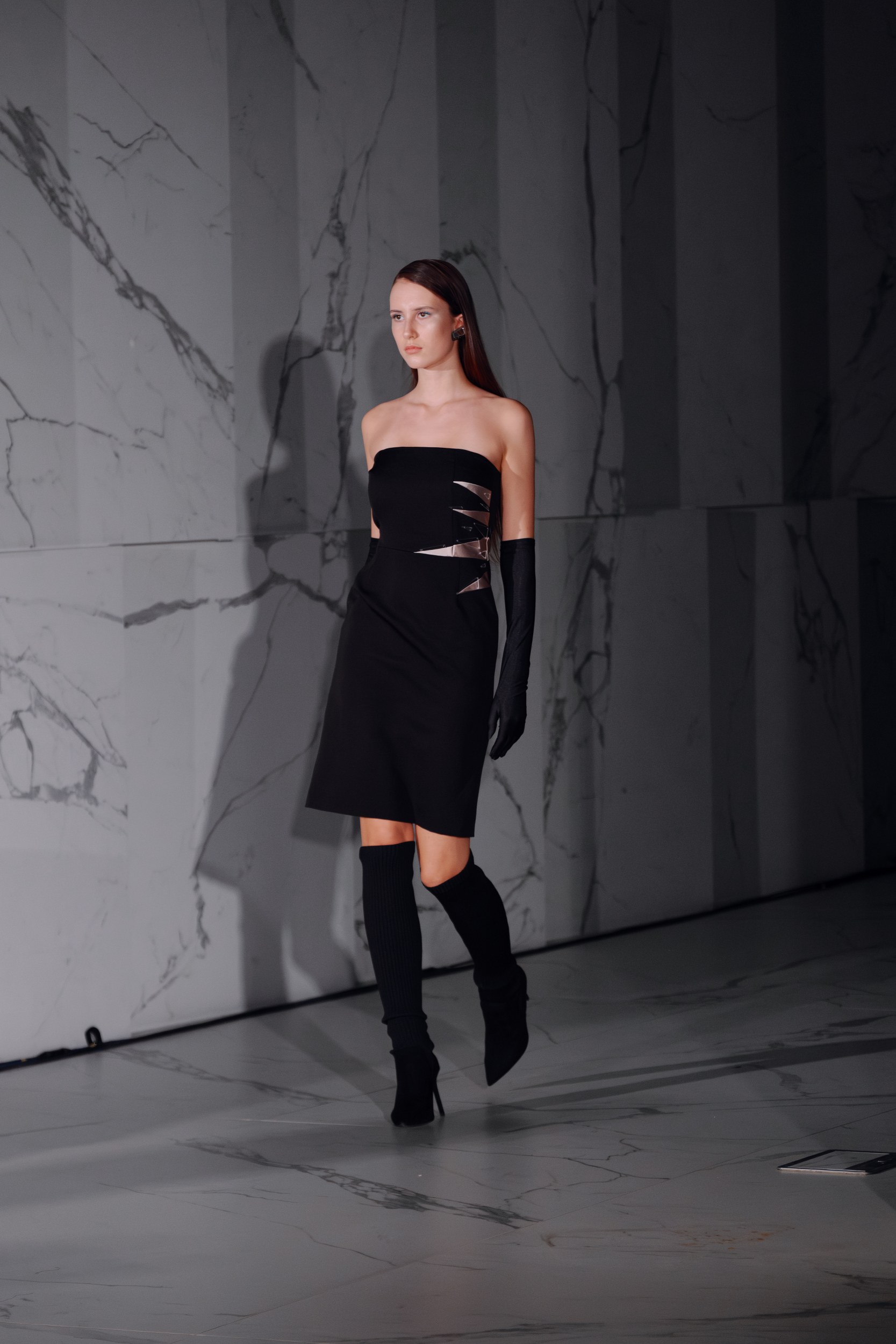 Model in black strapless dress on runway with marble backdrop.