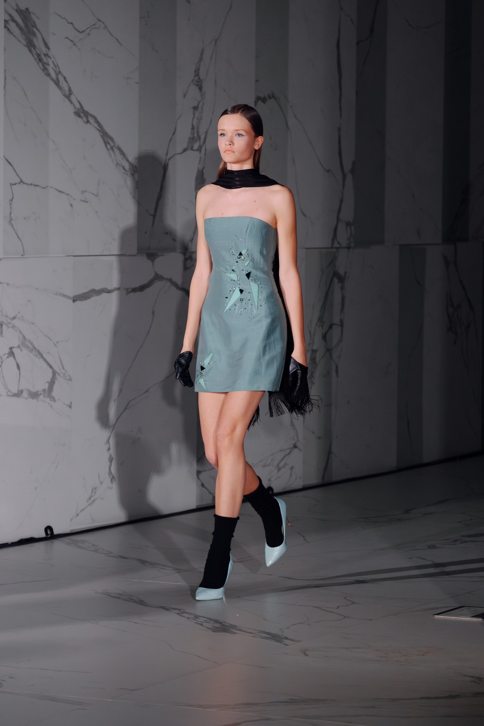 Model on runway in teal dress, marble background, black gloves and scarf. Fashion show.