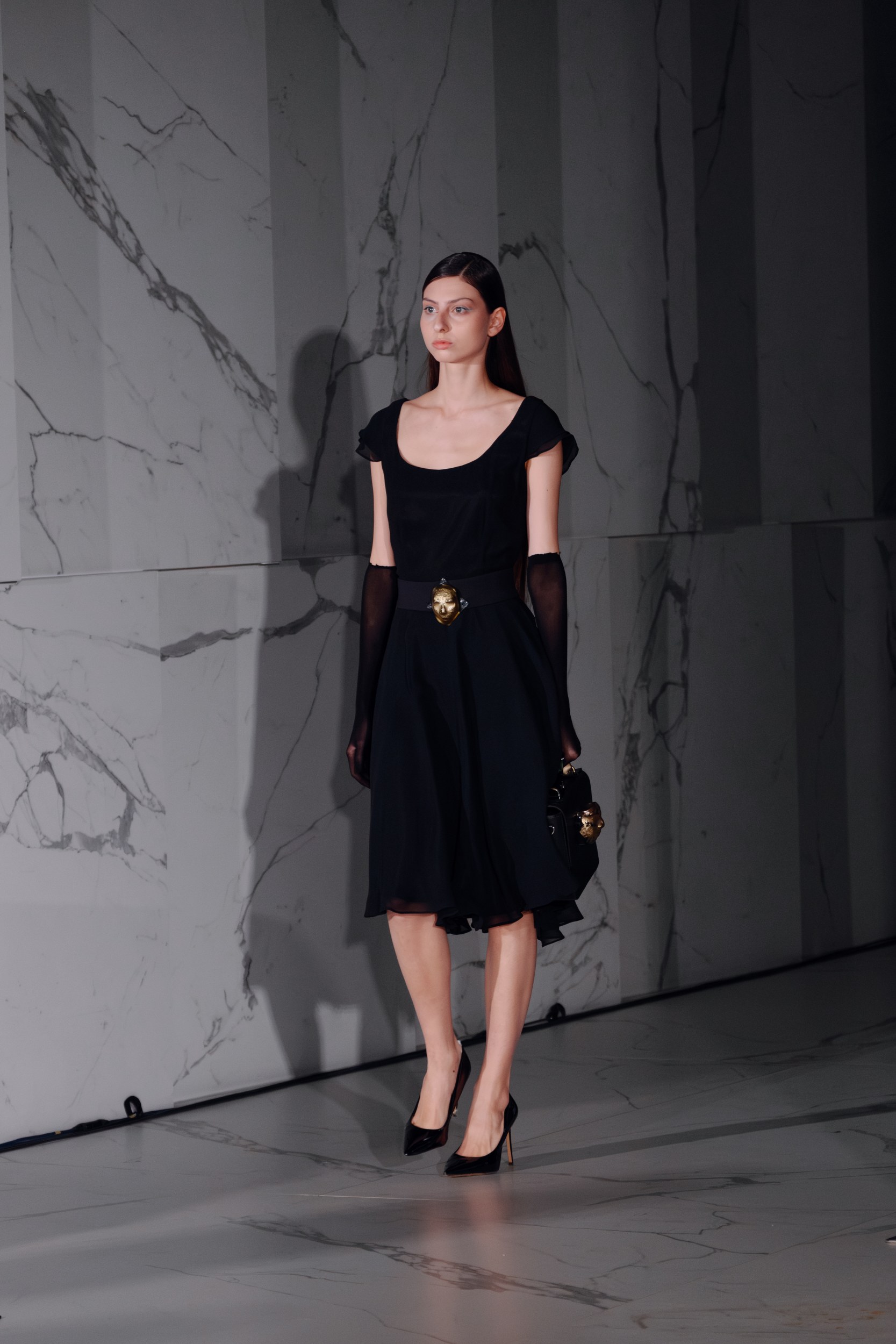 Model in elegant black dress walking on marble runway.