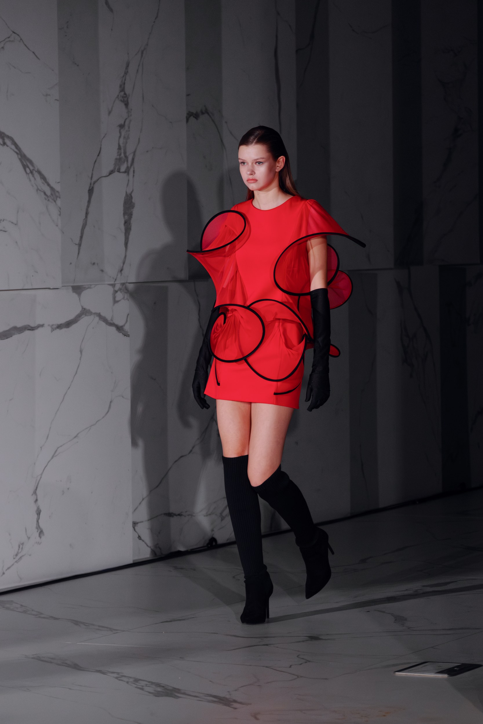 Model in red avant-garde dress on marble runway.