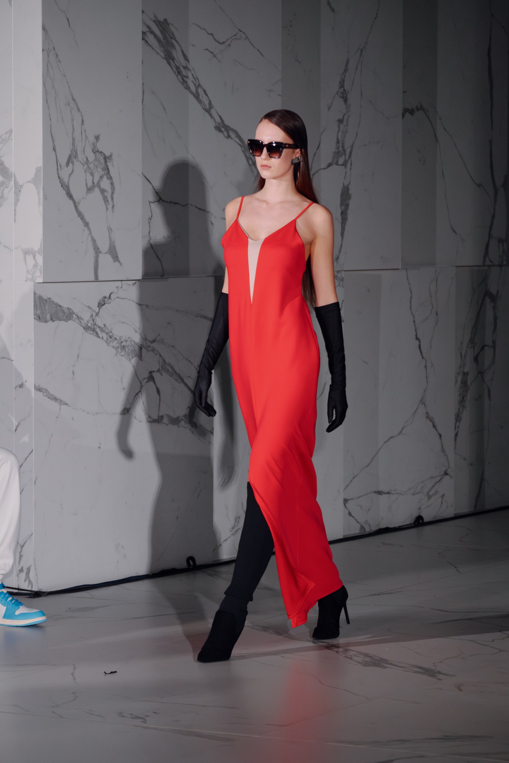 Model in red dress with black gloves on runway, marble backdrop.