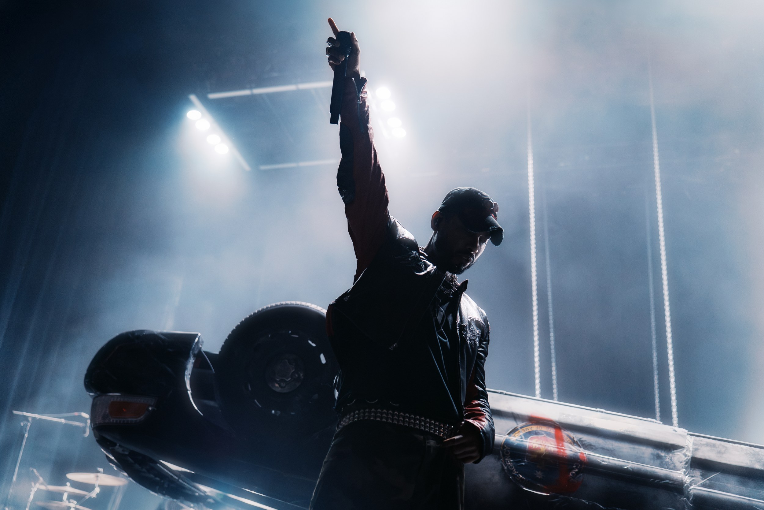 Performer on stage with raised arm, dim lights, overturned car in background.
