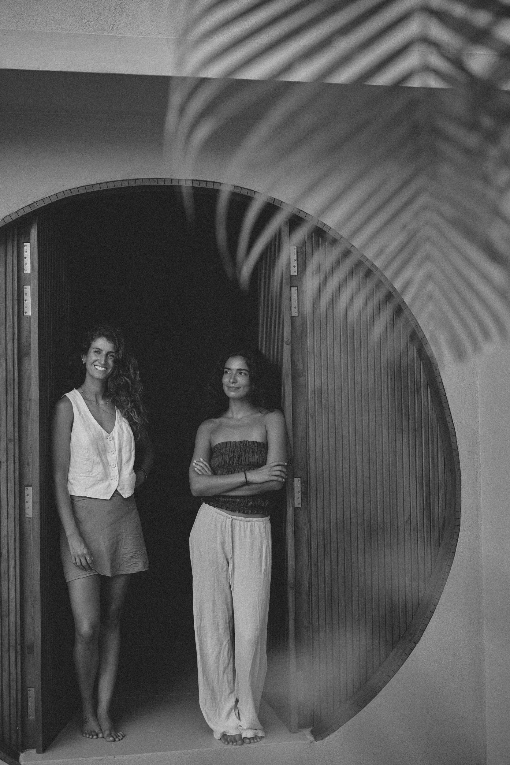 Two women smiling by a round wooden doorway, with palm leaf shadow above.
