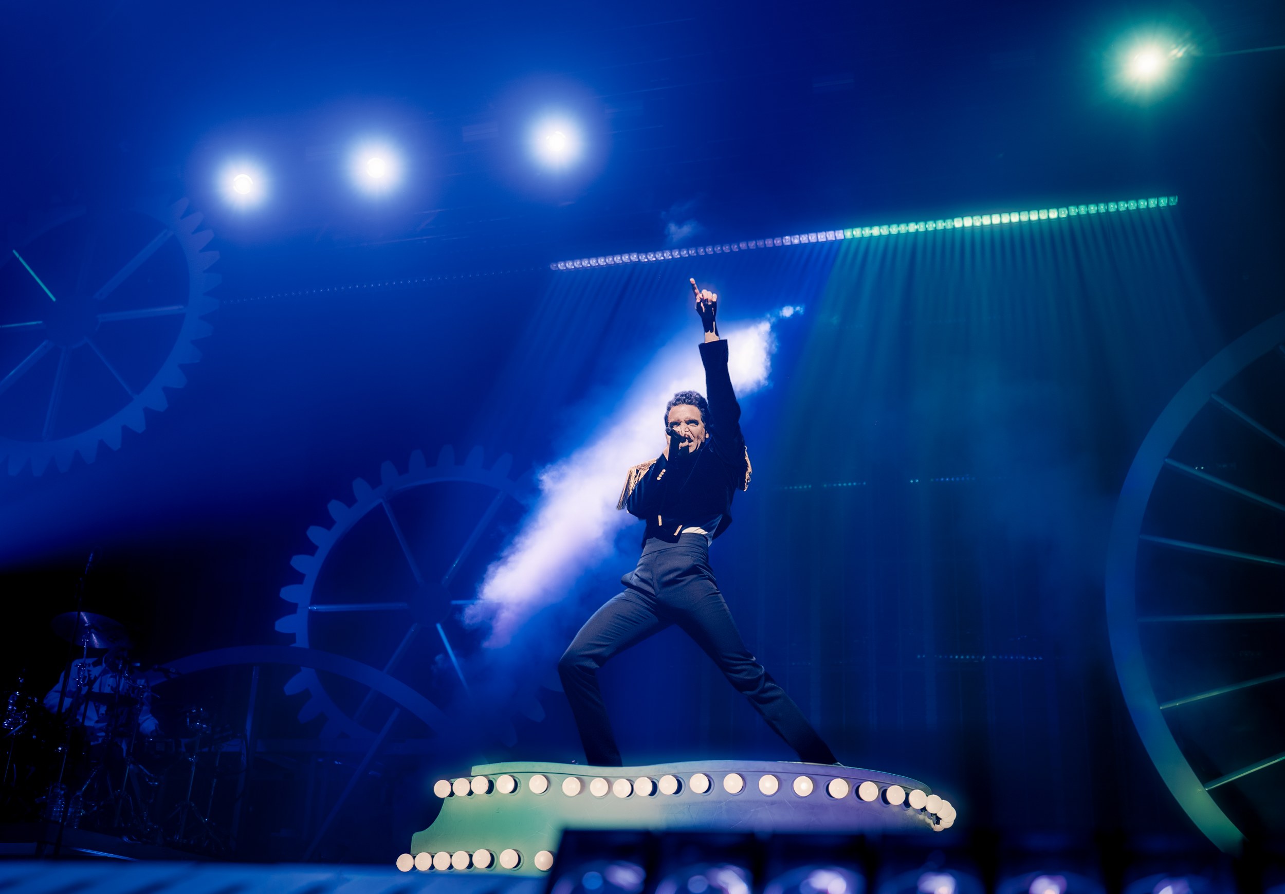 Performer on stage with dramatic lighting and gear backdrop, pointing upwards.