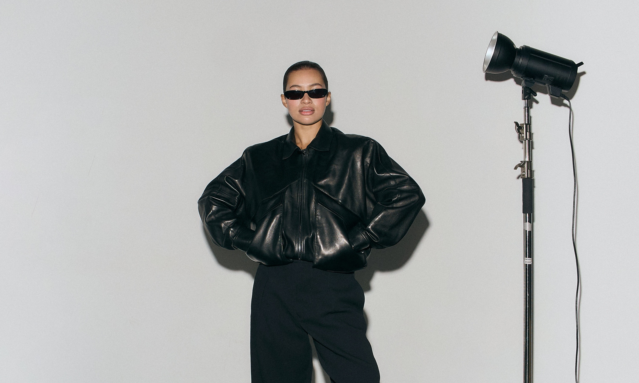 Fashion model wearing a black leather jacket and sunglasses in a studio setting.