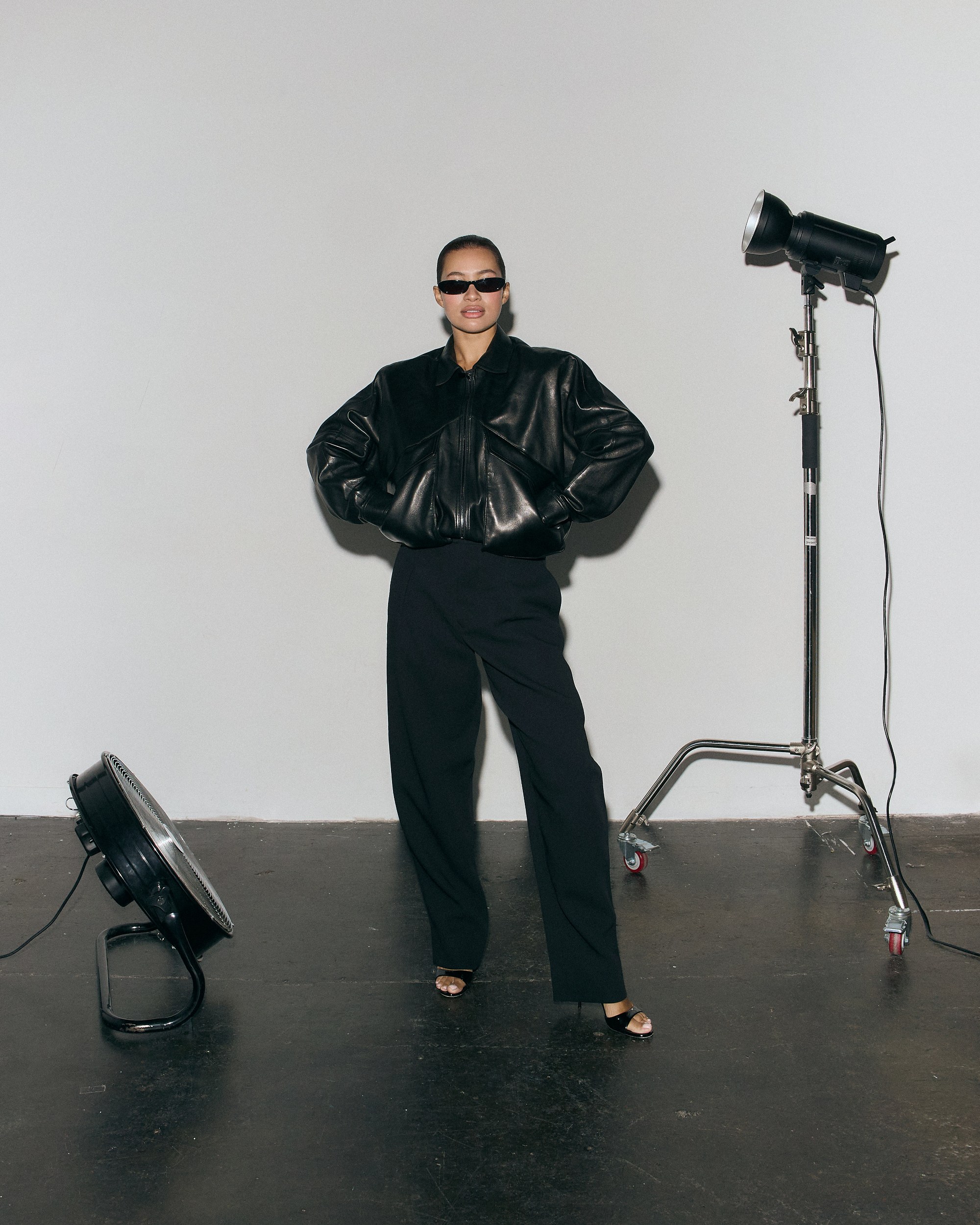 Person in black leather jacket, sunglasses, and pants, standing in a studio setting.