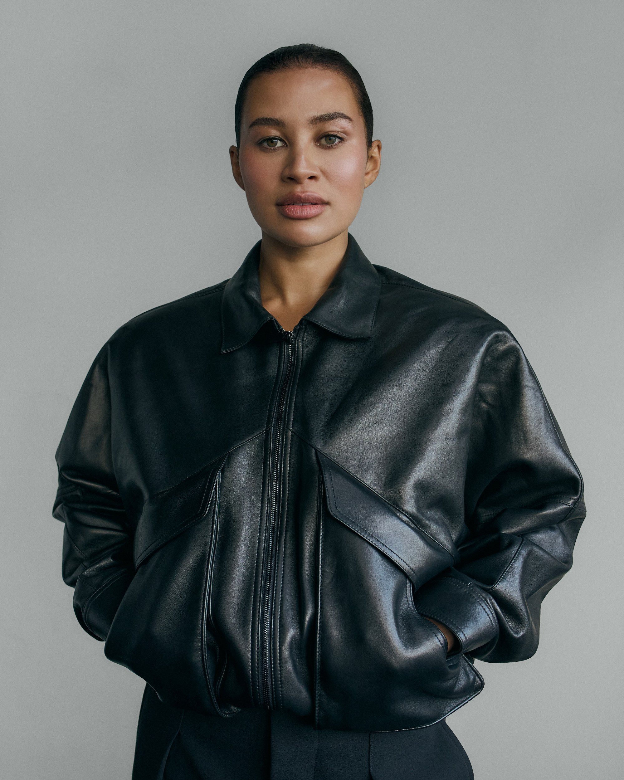 Woman in sleek black leather jacket with hands in pockets, neutral background.