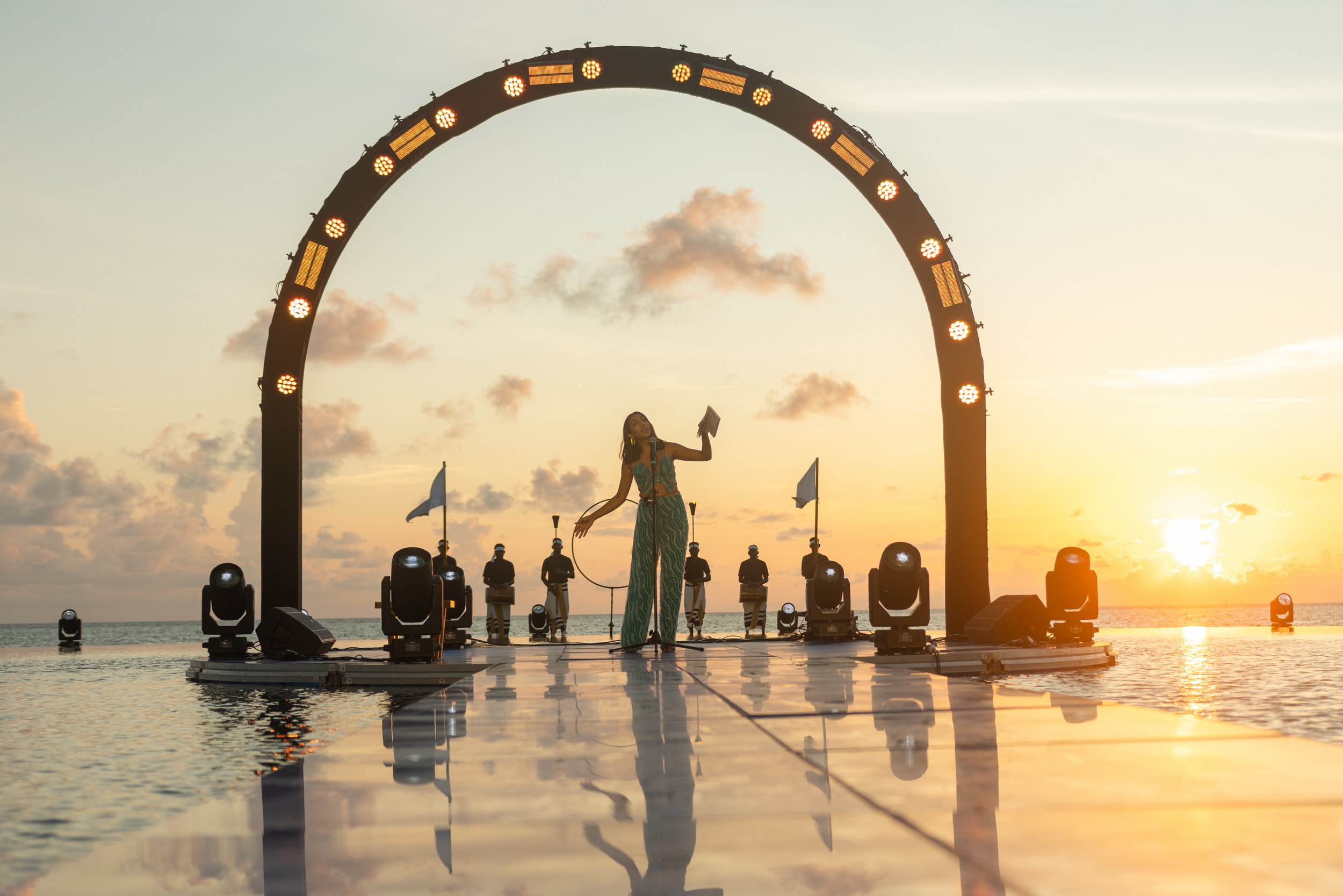 Sunset performance on a stage over water, lit archway, reflections, and audience.