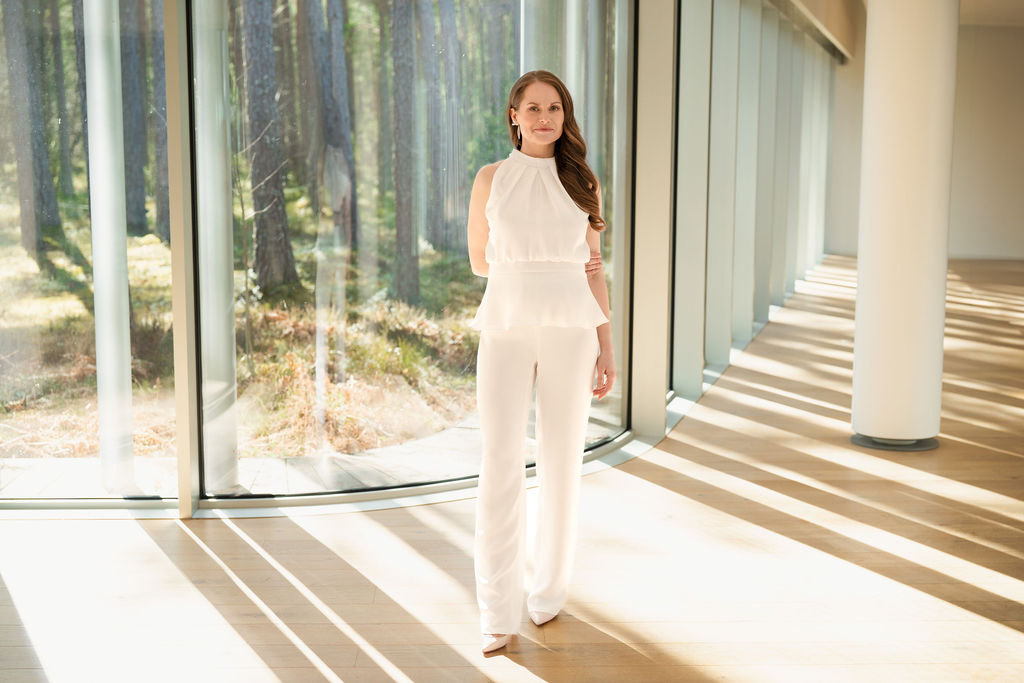 Woman in white outfit by large windows in a sunlit modern room.