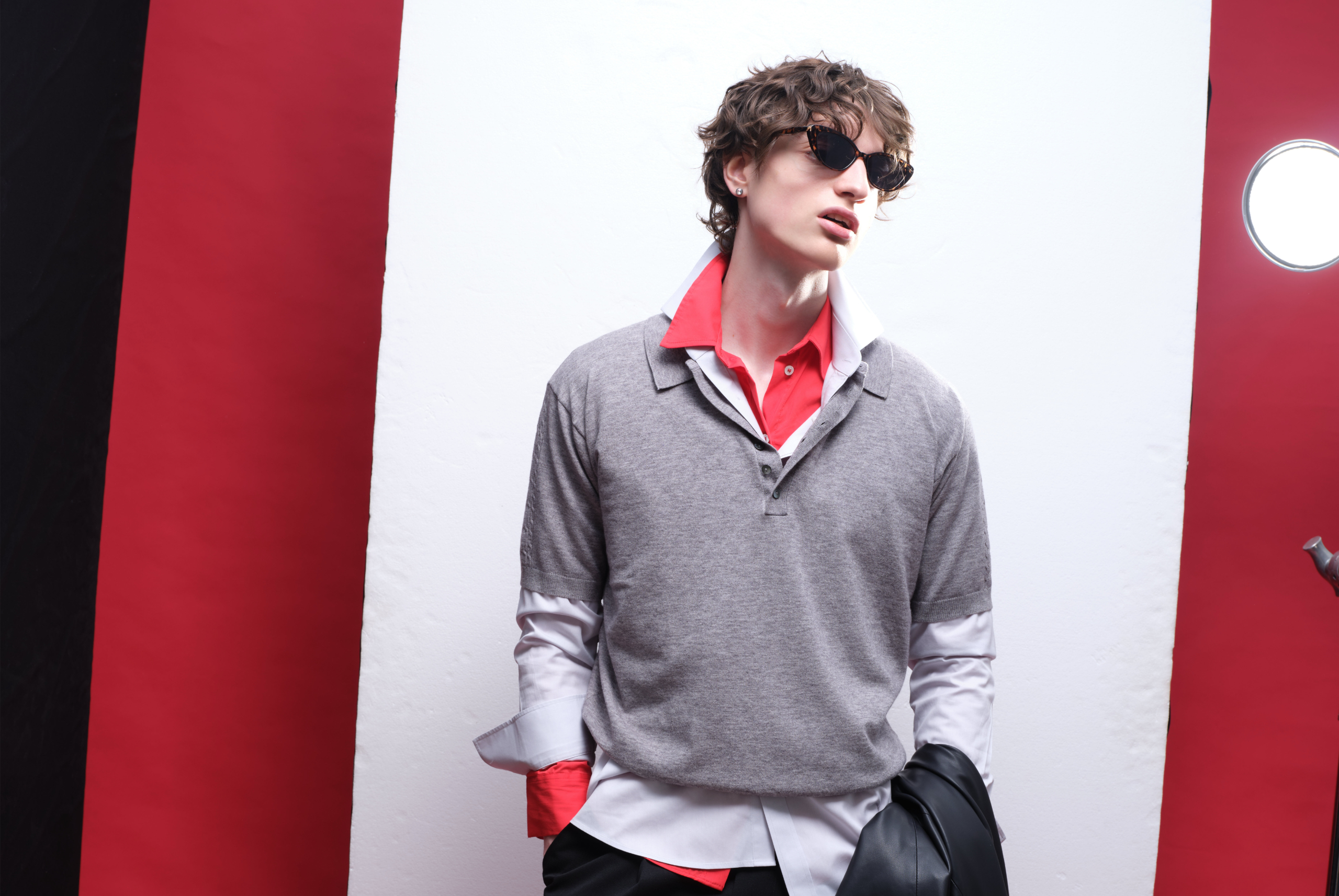 Fashionable model in layered outfit with sunglasses against red and white backdrop.