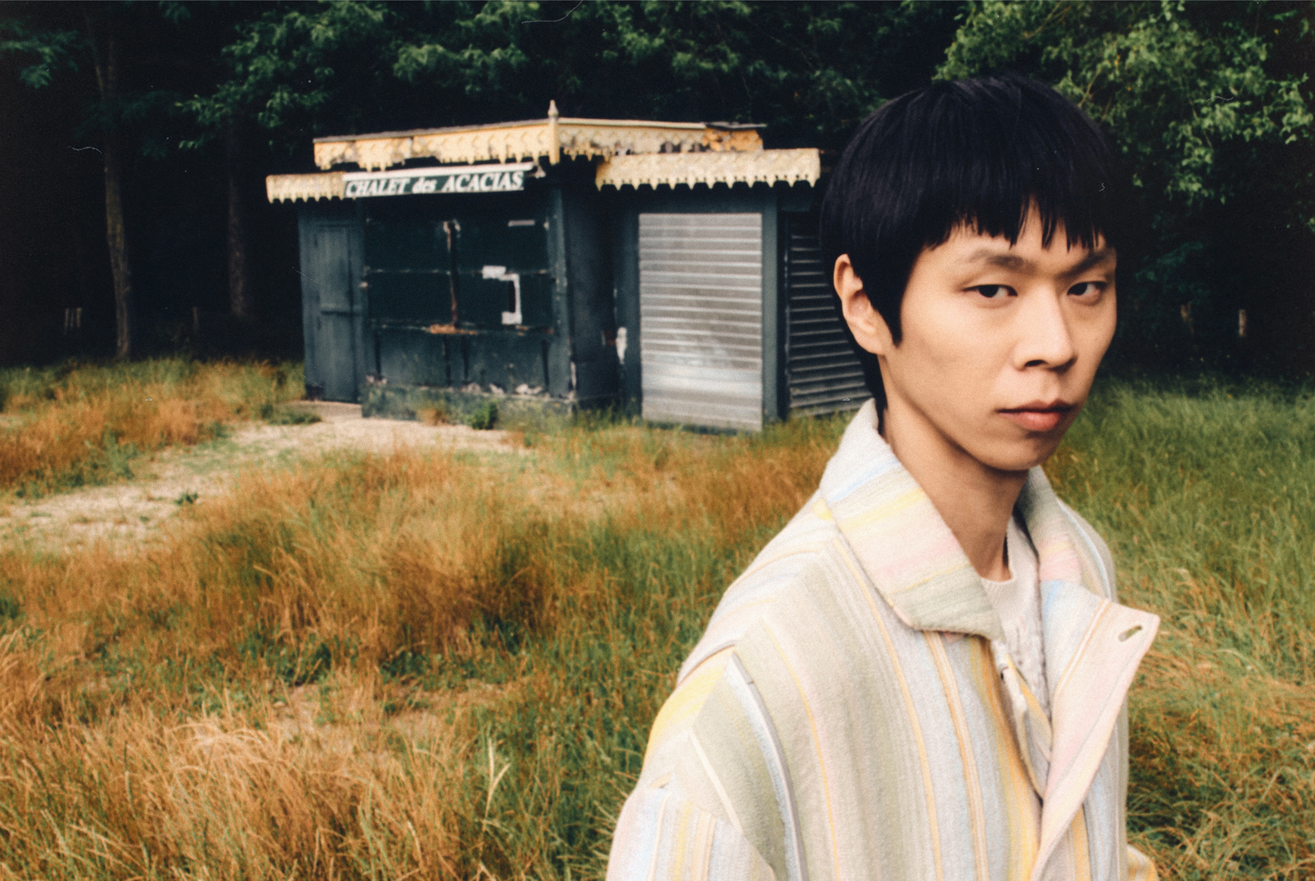 Person standing by an old building in grassy field with trees behind.