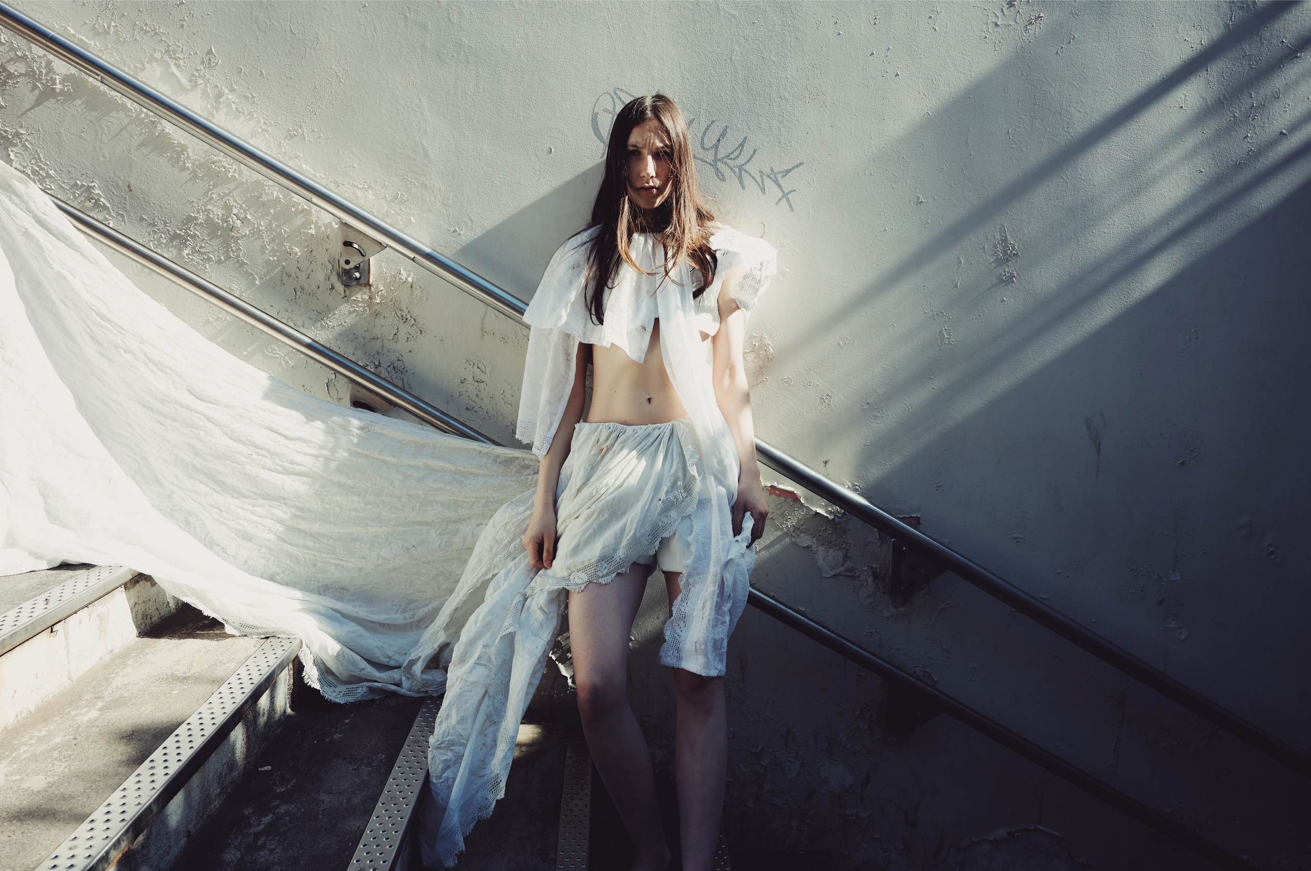 Woman in ethereal white dress standing on sunlit stairs.