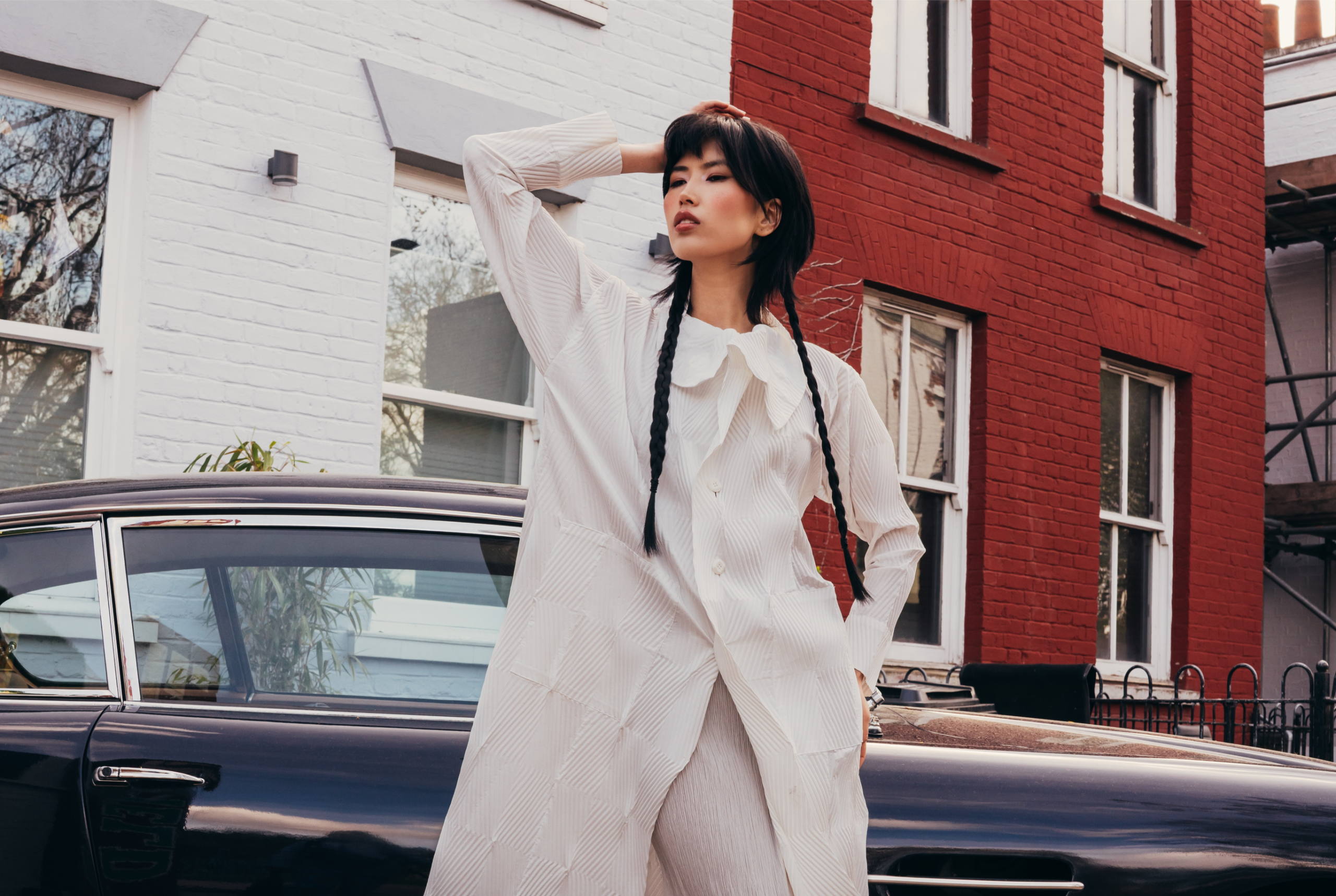 Fashionable person in white outfit leaning by vintage car outside colorful building.