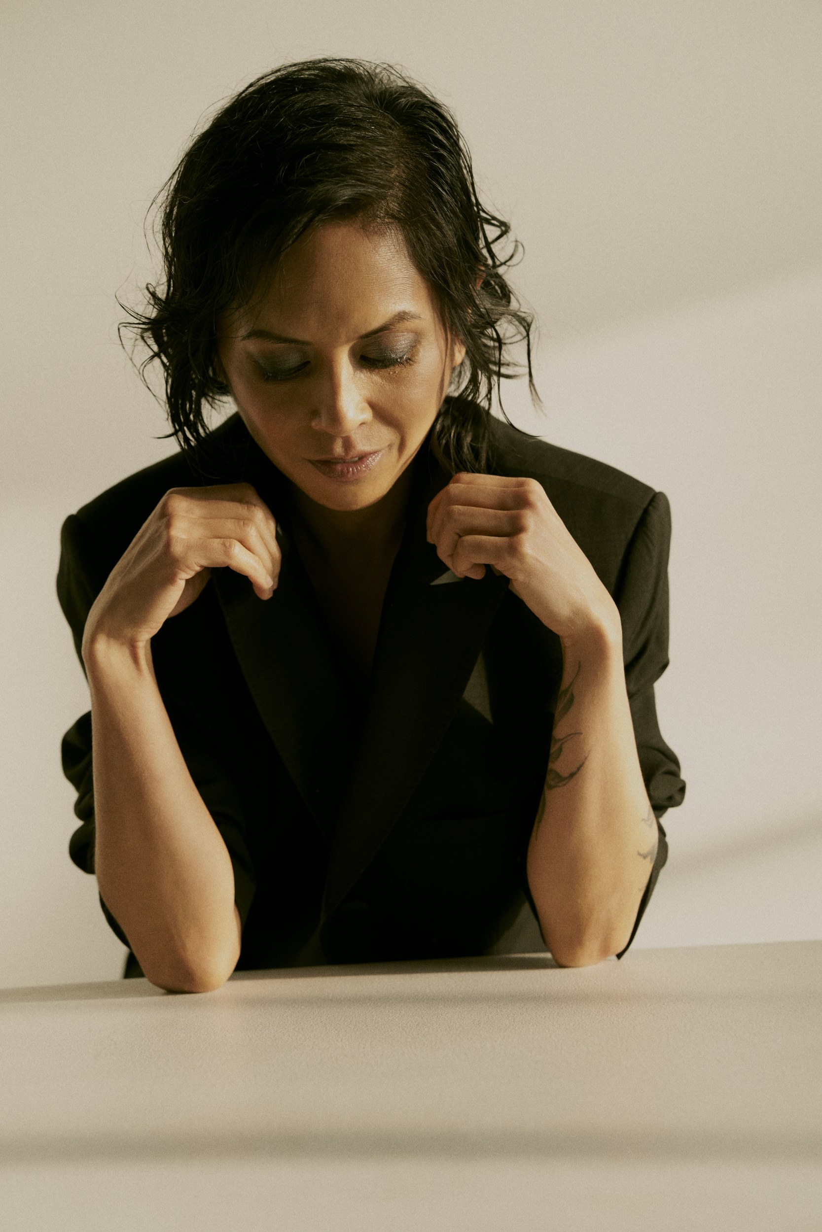 Pensive person in a black suit with tattooed arms leaning on a table.