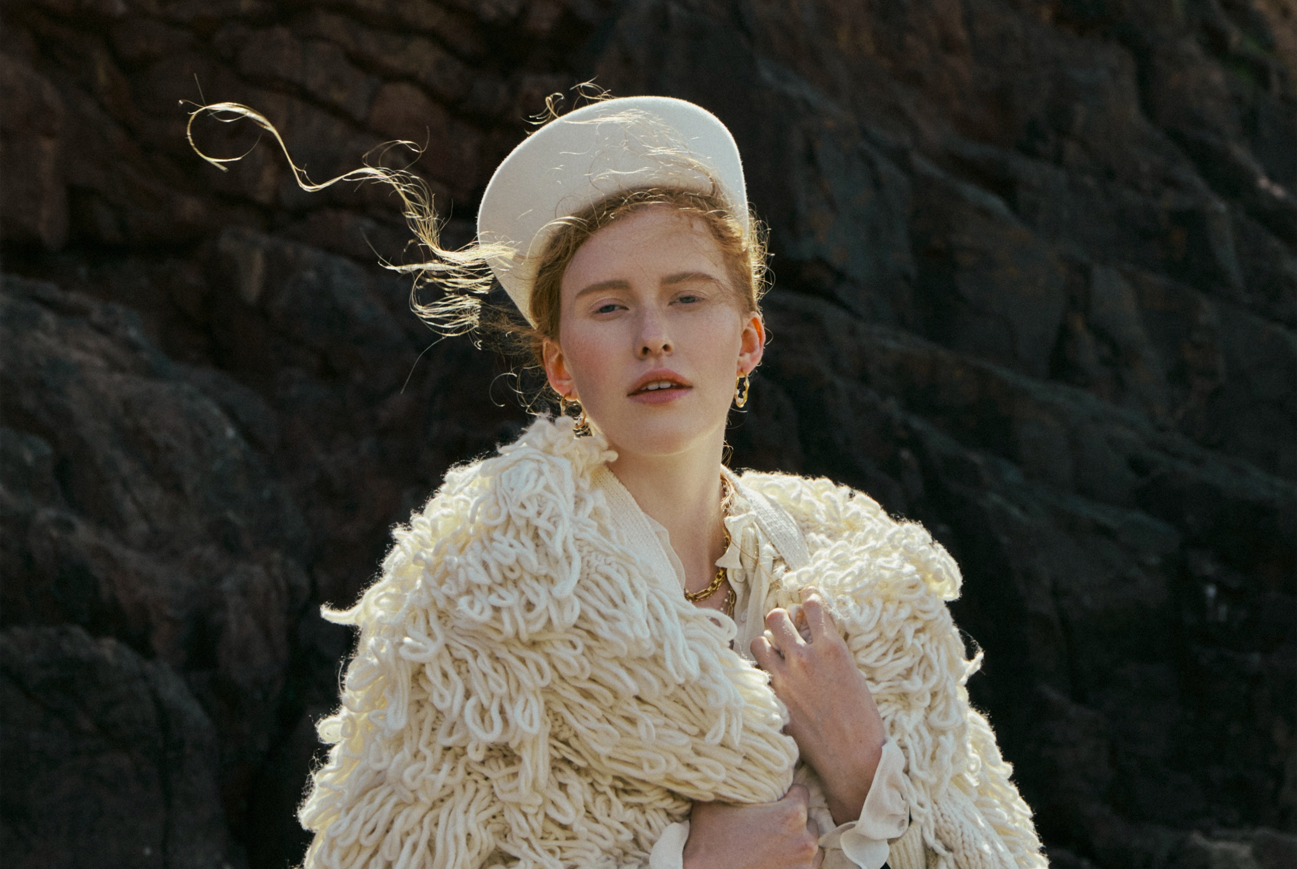 Person in textured white outfit with hat standing against rocky background.